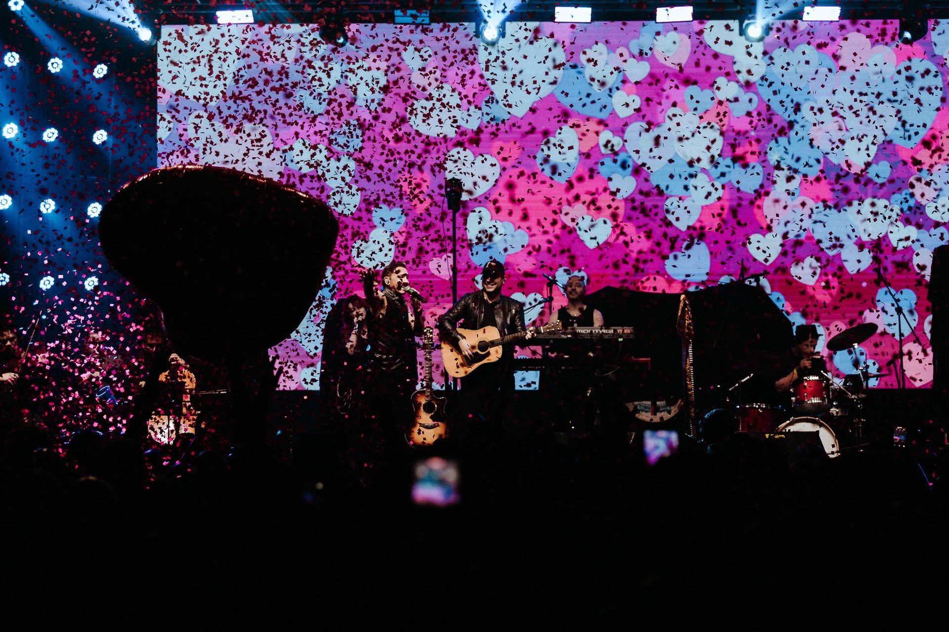 Banda se apresentando no palco com confetes caindo em frente a uma tela colorida.