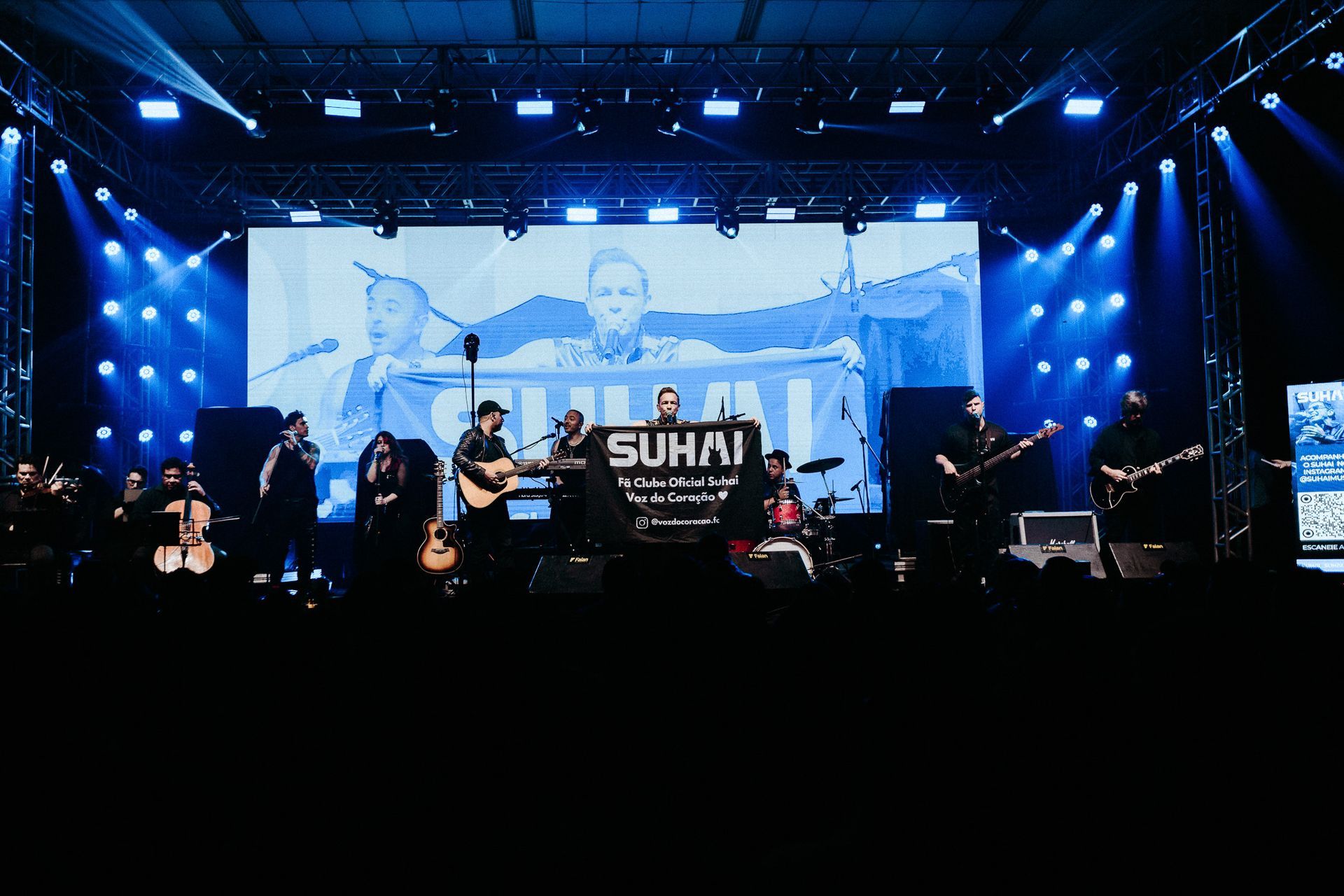 Banda se apresentando no palco com luzes azuis e um telão ao fundo exibindo o nome da banda.