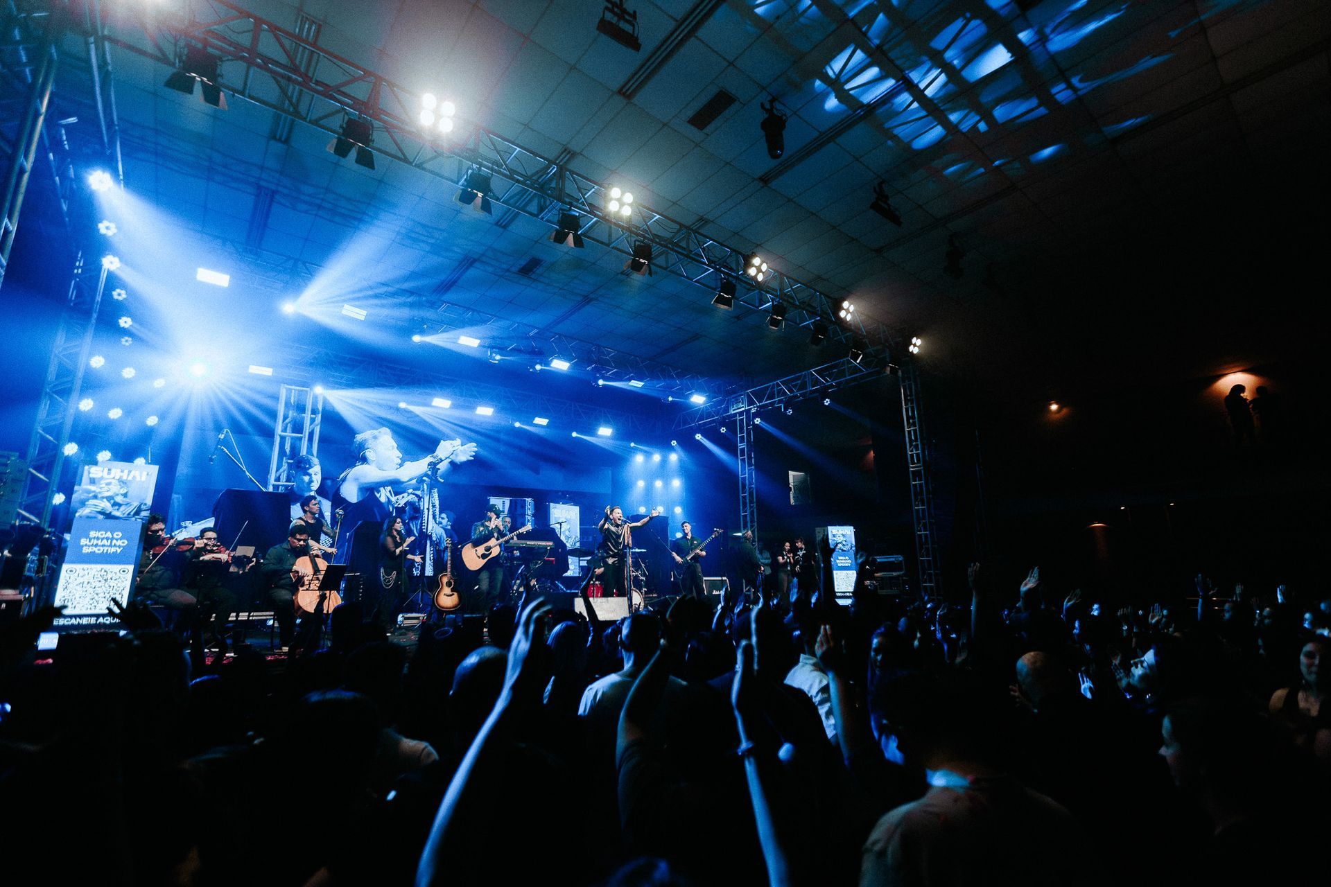 Banda se apresentando no palco, luzes azuis brilhantes, público com as mãos erguidas, local fechado.