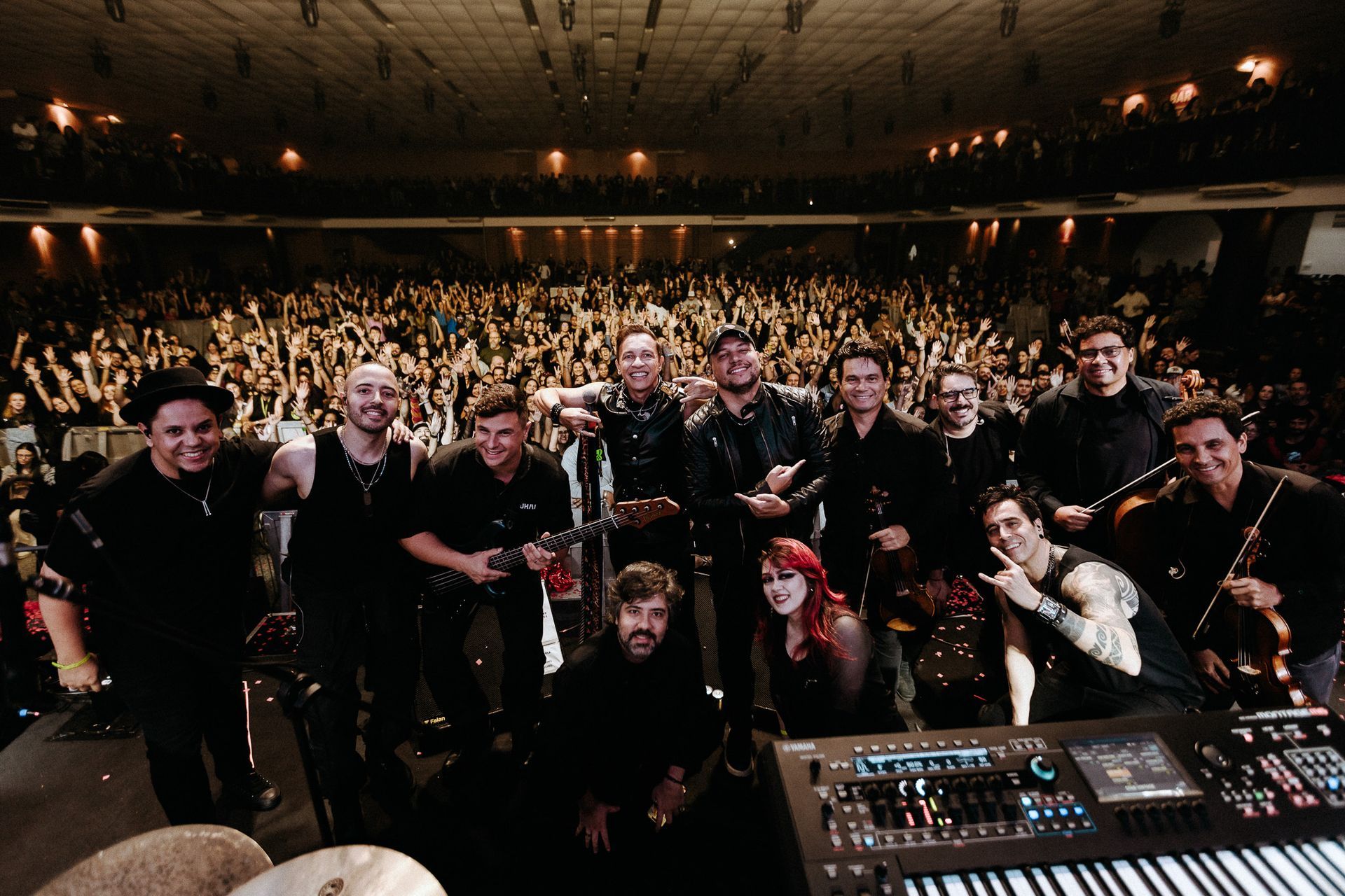 Banda no palco com grande público em uma sala de concertos, todos de frente para a câmera, sorrindo.