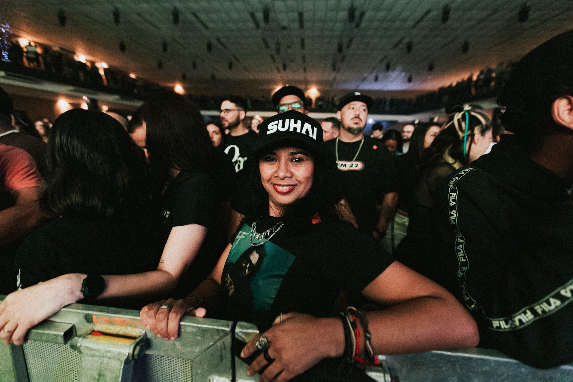 Mulher sorrindo em um show, usando chapéu e pulseiras, encostada em uma grade. Muitas pessoas ao fundo.