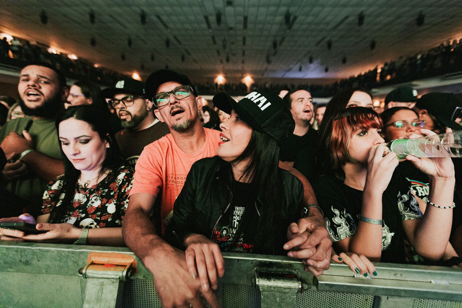Público em um show: Pessoas em um local de espetáculo, encostadas em uma grade. Algumas cantam, bebem e olham para cima. 