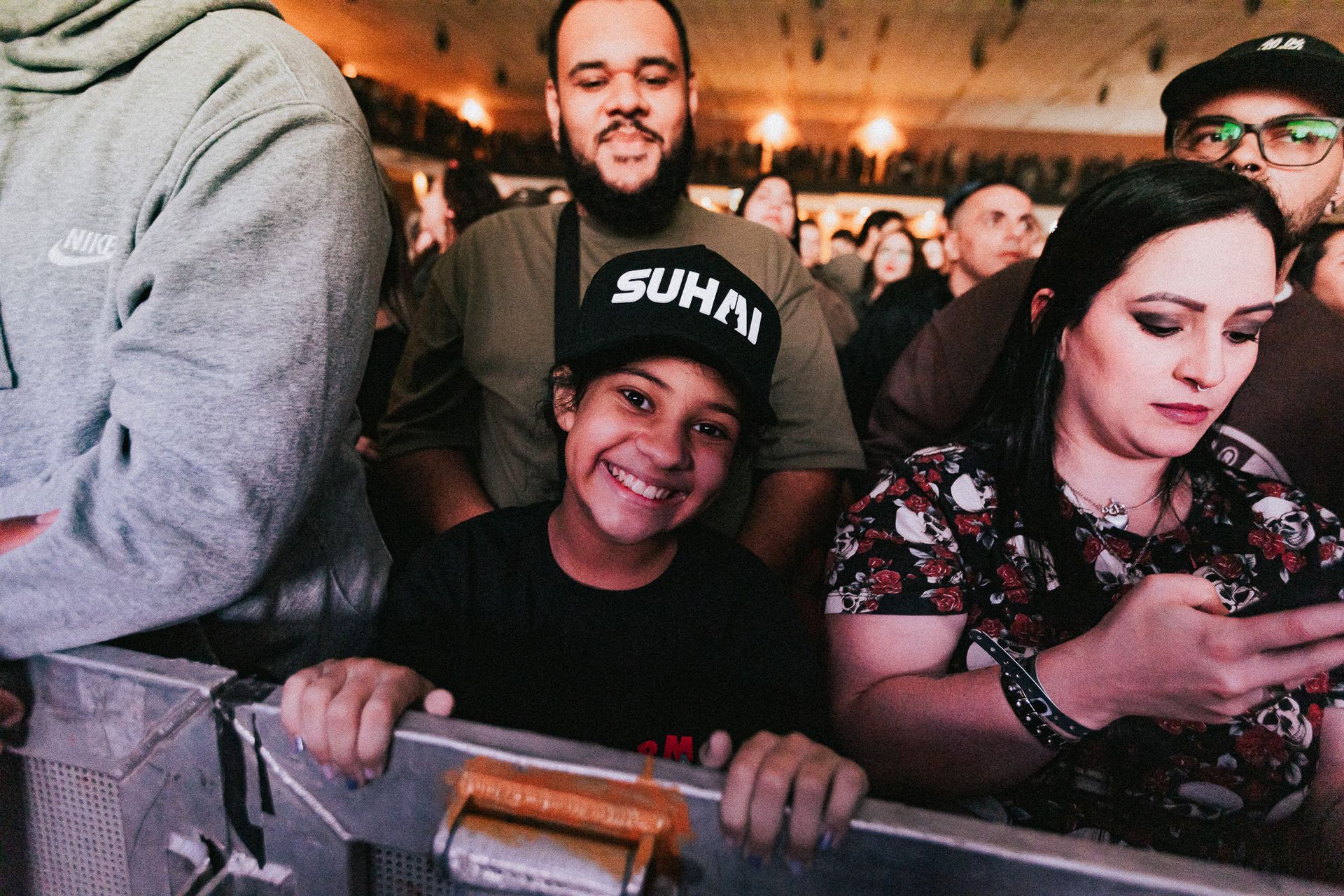 Uma pessoa jovem sorri para a câmera, encostada em uma grade de metal em um show. Multidão ao fundo.