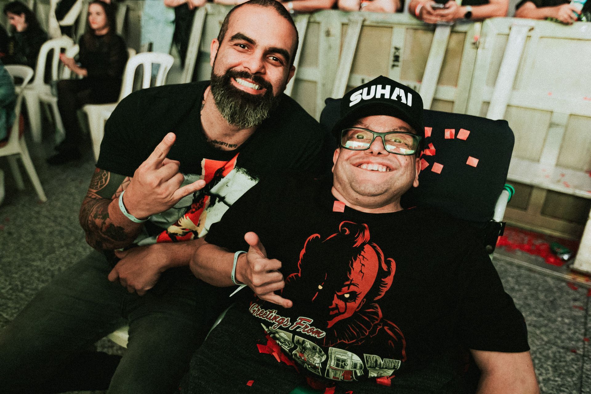 Dois homens sorrindo, um em uma cadeira de rodas, com o outro braço levantado e ambos vestindo camisetas escuras.