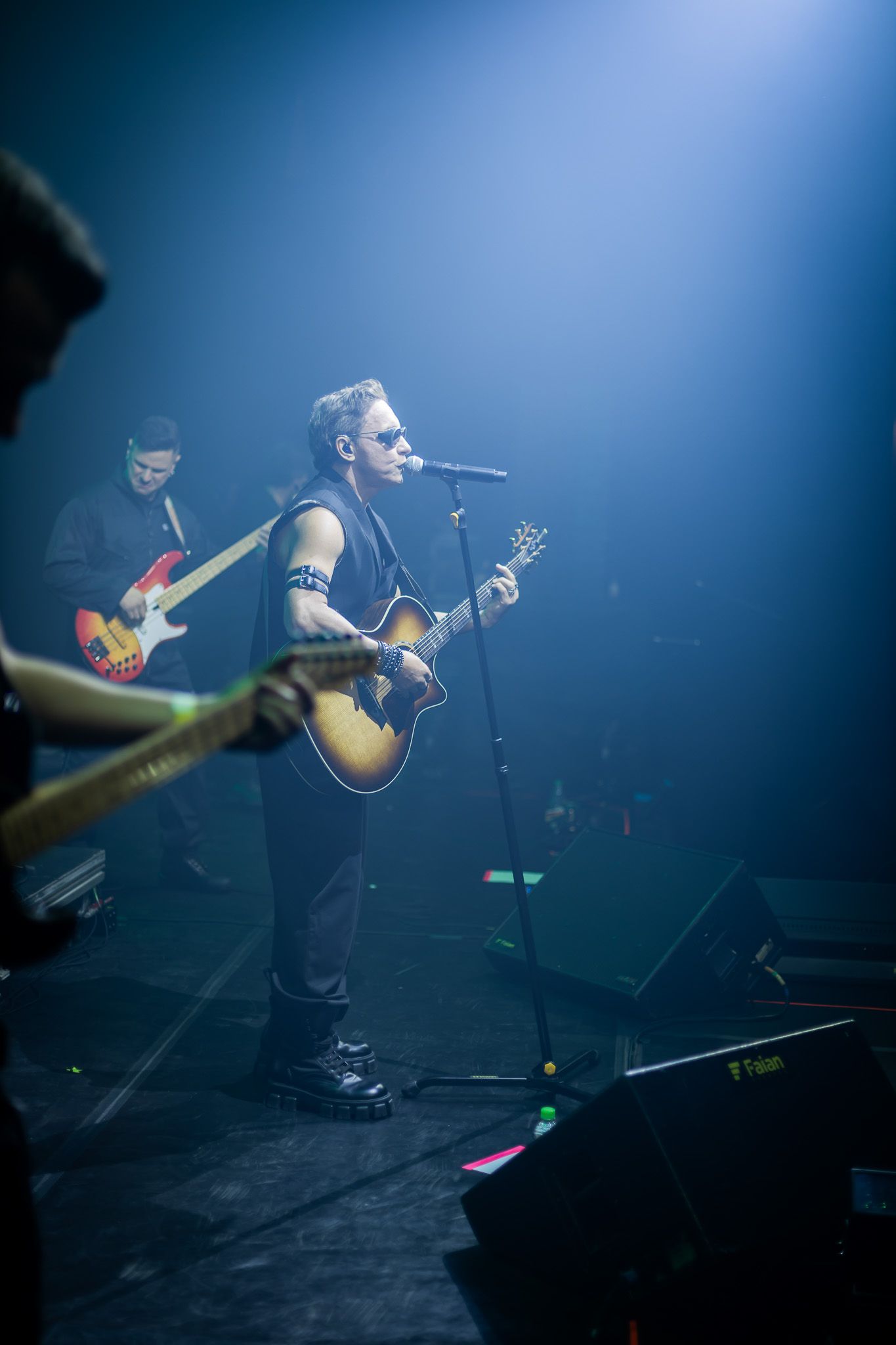 Músicos se apresentam em um palco de concertos, sob um holofote azul que ilumina o cantor central que toca violão.