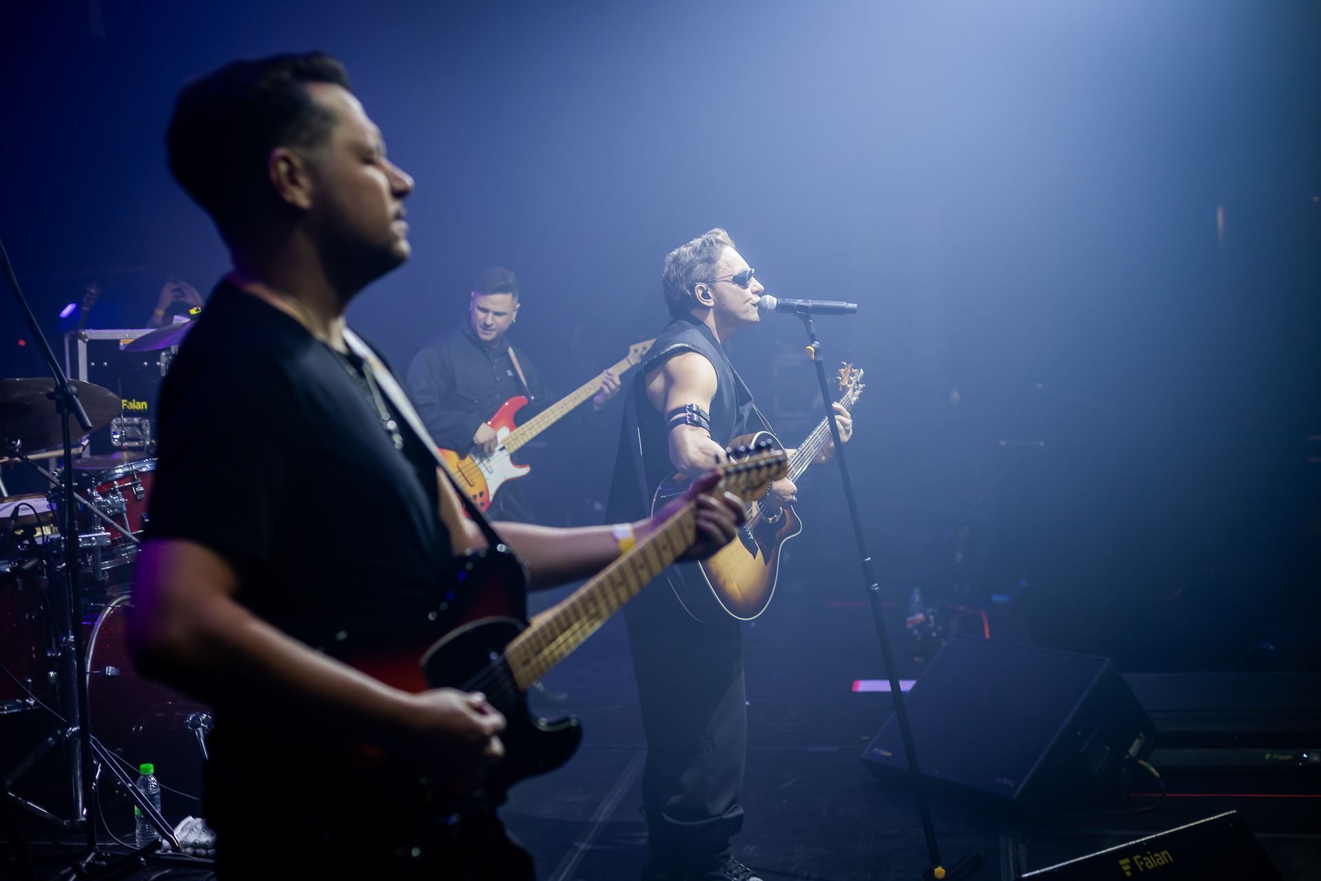 Uma banda se apresenta em um palco pouco iluminado, com um guitarrista e um cantor de óculos escuros ao microfone.