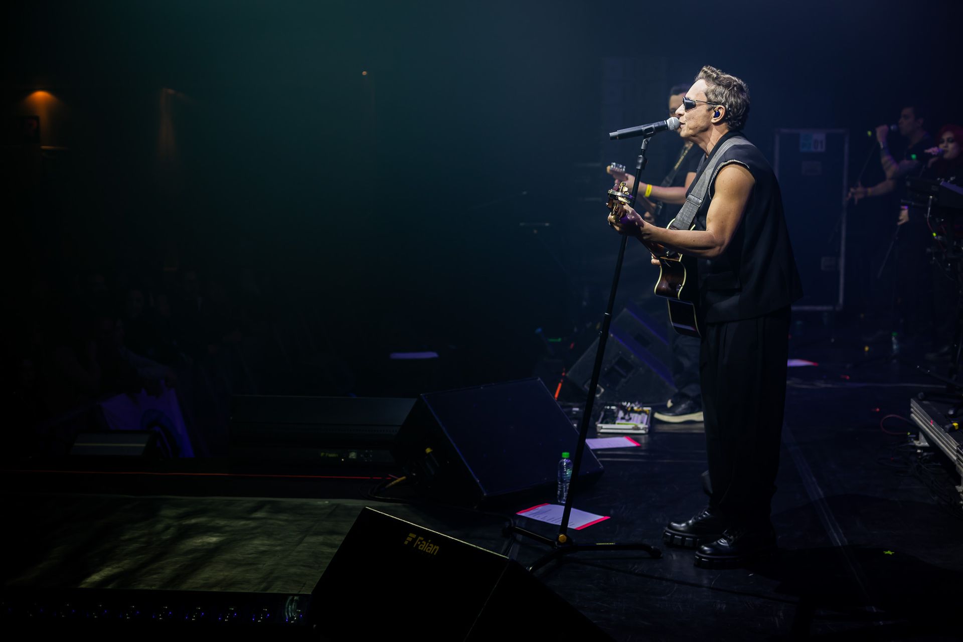 Um músico se apresenta em um palco com pouca luz, cantando ao microfone enquanto toca guitarra elétrica.