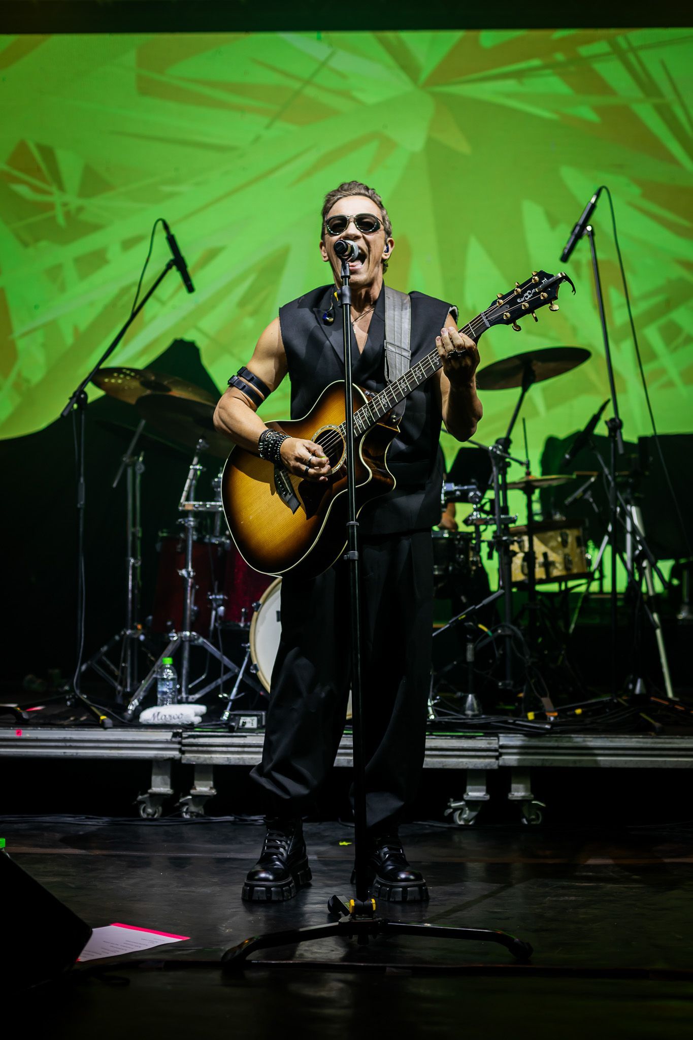 Um músico tocando violão e cantando ao microfone em um palco com um fundo geométrico verde brilhante.