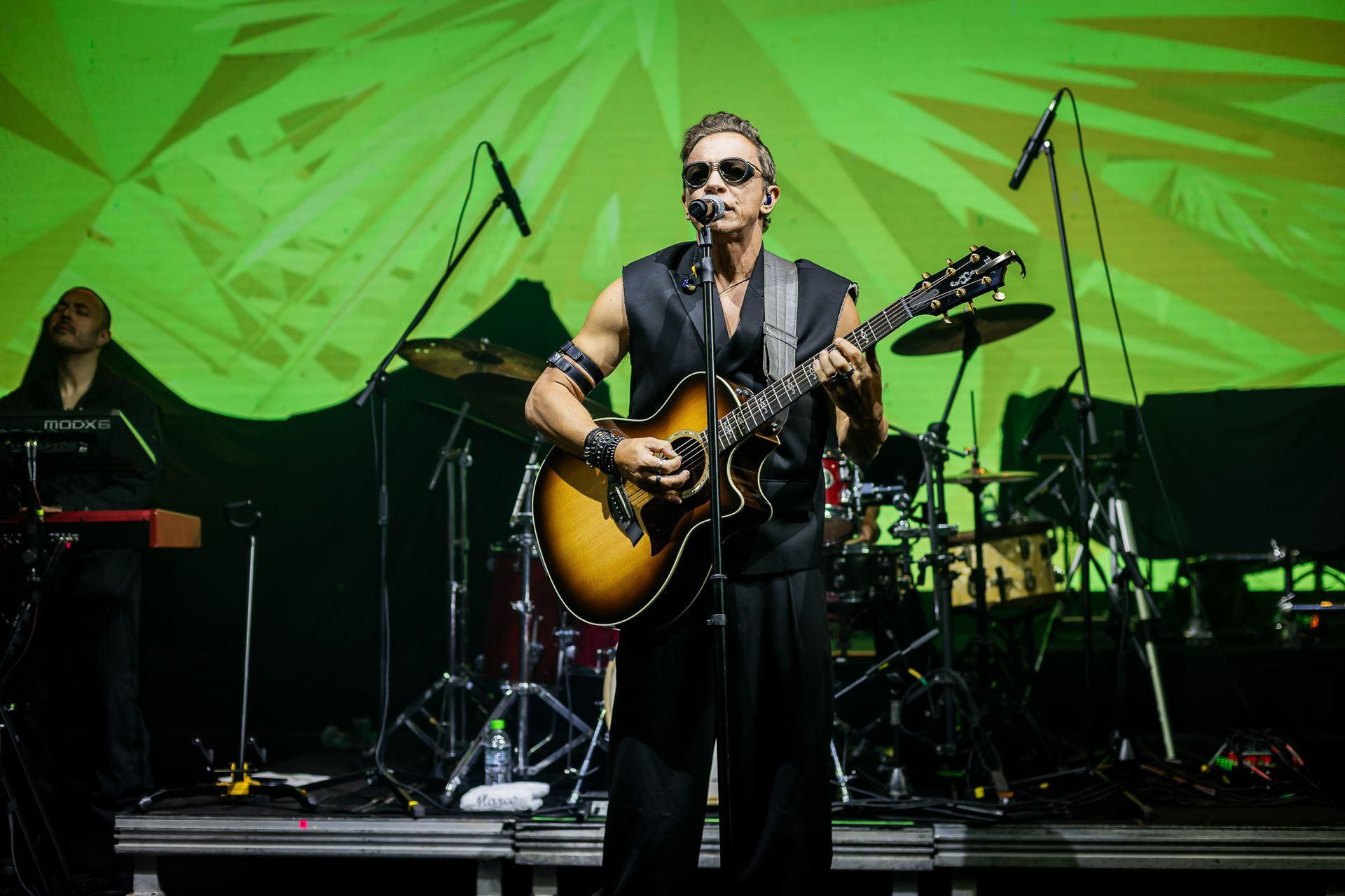 Um artista vestido com um traje preto sem mangas toca violão no palco contra um fundo texturizado verde vibrante.