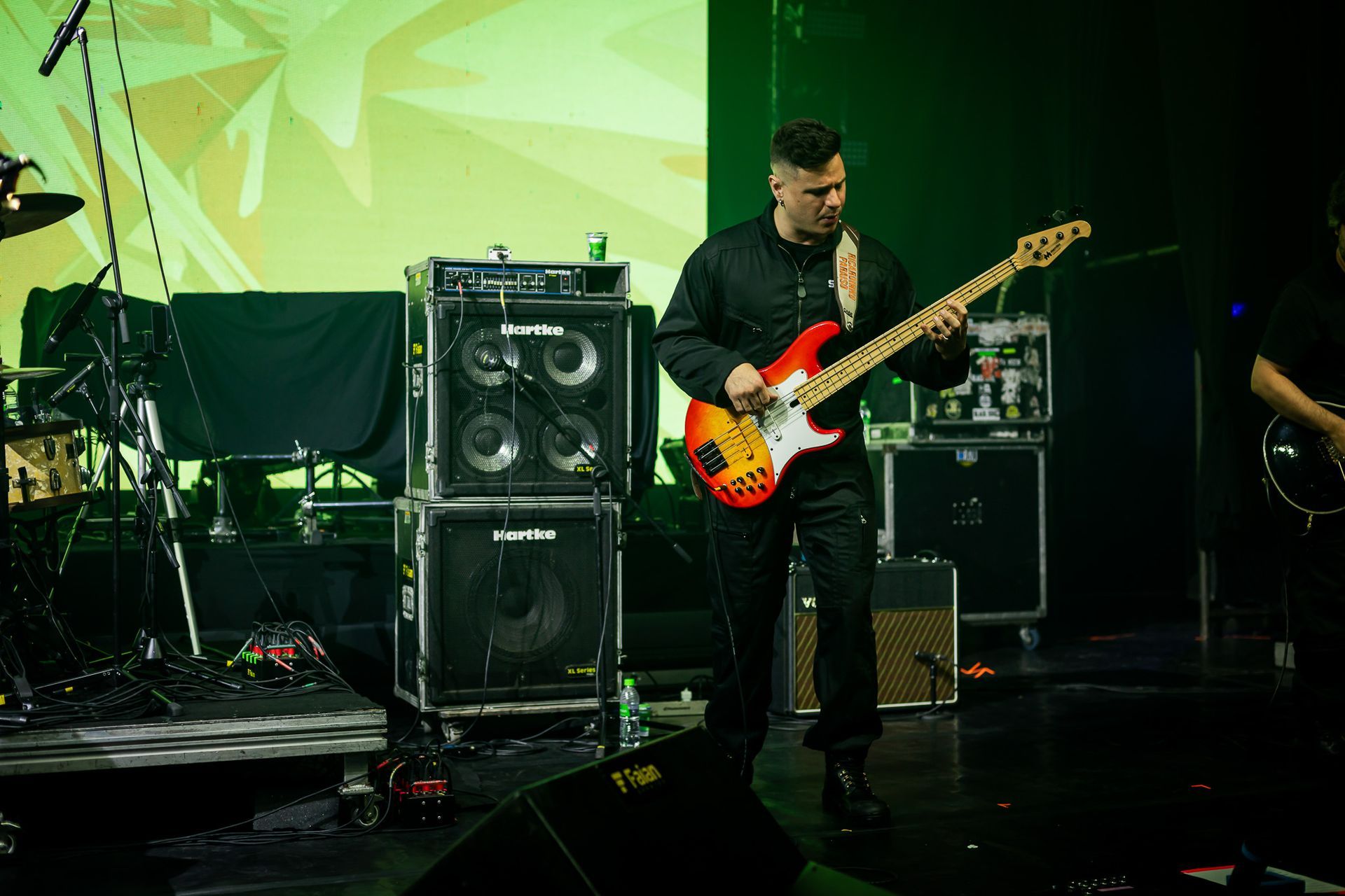 Um músico toca um baixo elétrico em um palco escuro, em frente a grandes caixas de som pretas e um fundo verde.