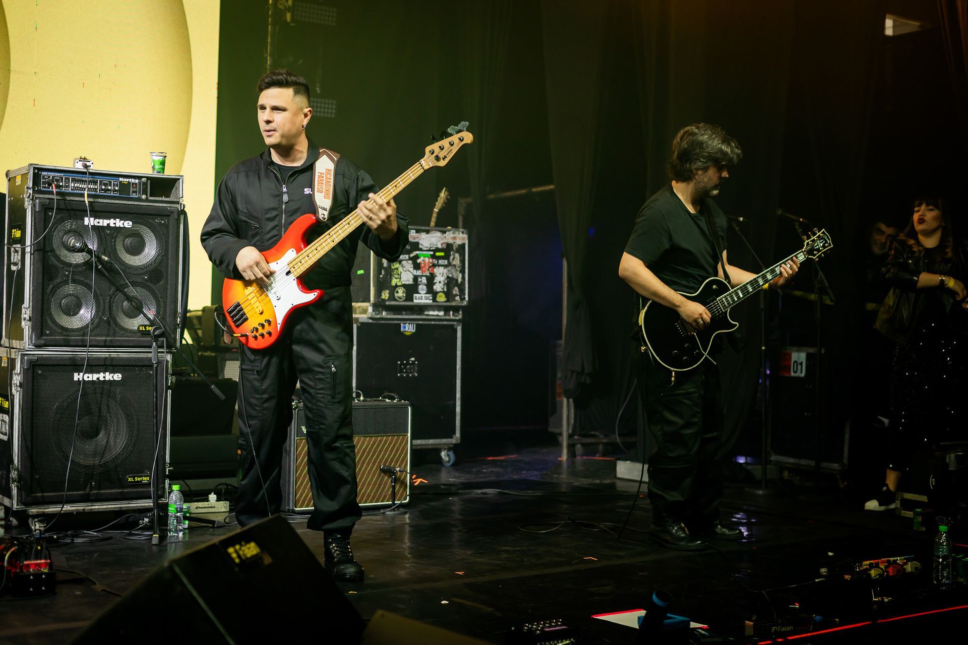 Dois músicos se apresentam no palco com guitarras elétricas e amplificadores sob iluminação fraca.