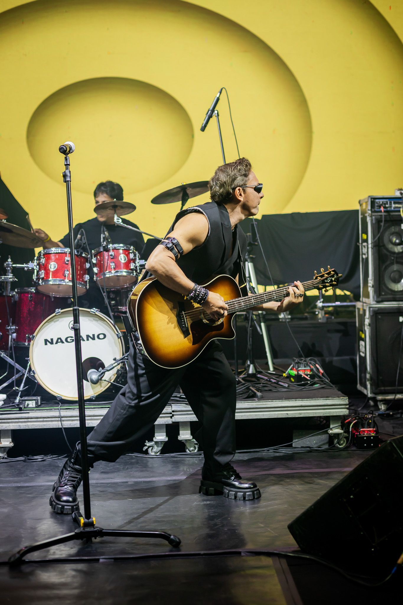 Um músico toca um violão eletroacústico no palco, em frente a um baterista, tendo como pano de fundo um amarelo vibrante.