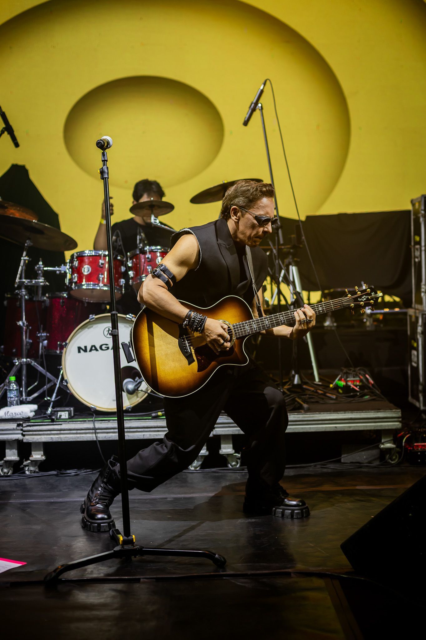 Um músico de colete preto toca uma guitarra elétrica sunburst em um palco com bateria contra um fundo amarelo.