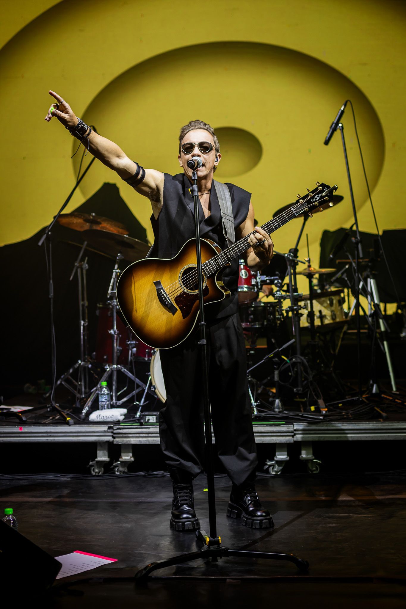 Um artista em um palco com um fundo circular amarelo, tocando violão e gesticulando com a mão direita.