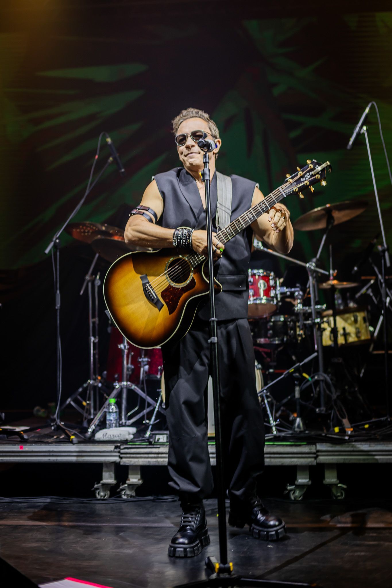 Um músico usando óculos escuros e um colete sem mangas toca violão em um palco com bateria ao fundo.