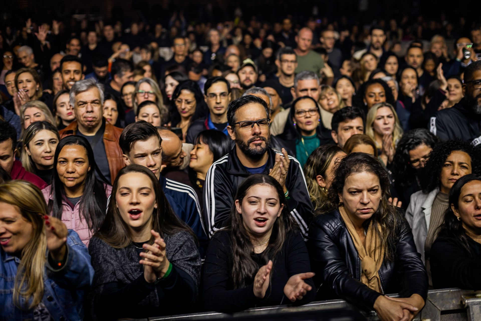 Uma multidão diversificada em um evento em local fechado, com pessoas em primeiro plano aplaudindo e olhando para o palco.