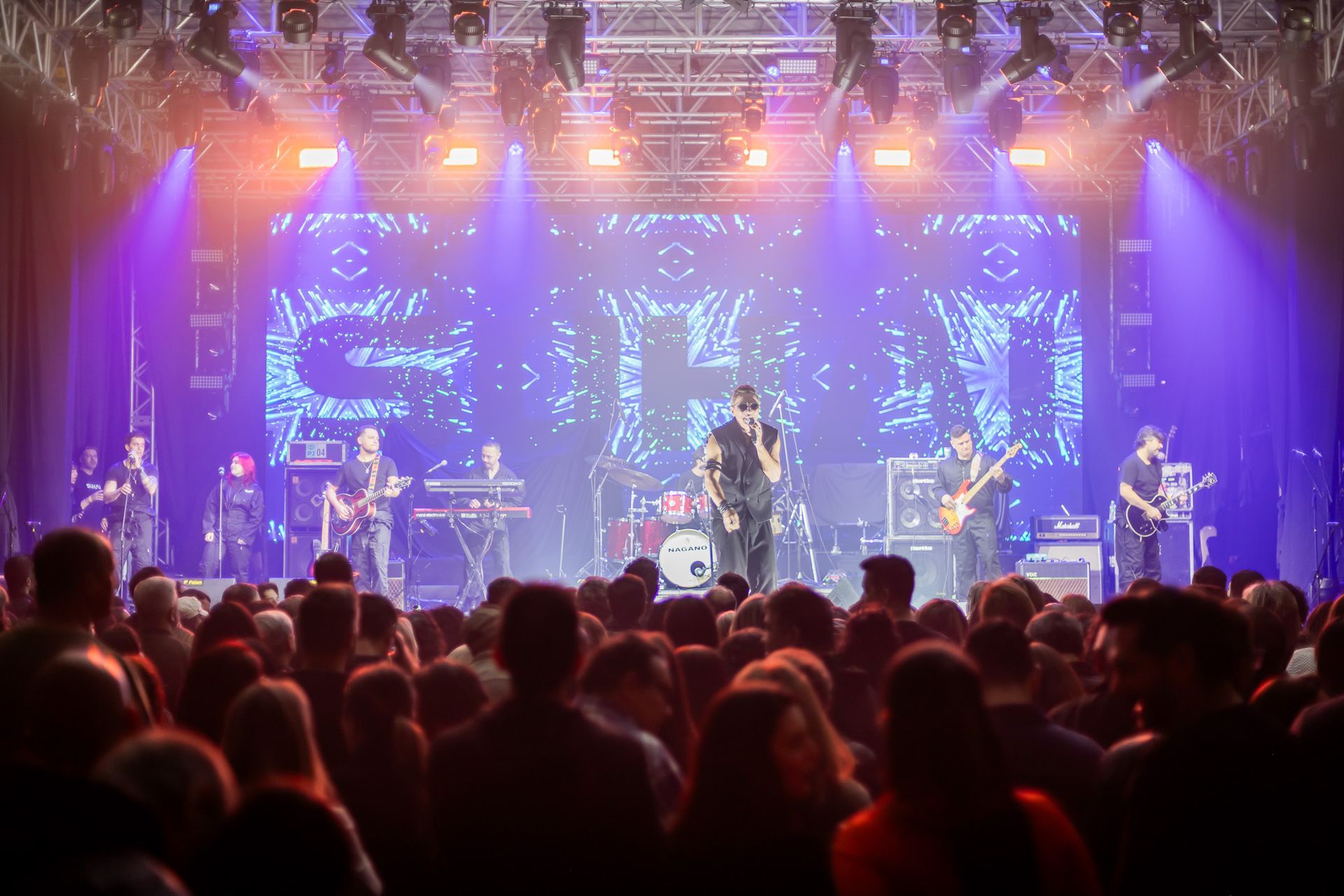 Uma banda se apresenta em um palco de shows bem iluminado, com um grande telão de LED ao fundo, para uma multidão de fãs.