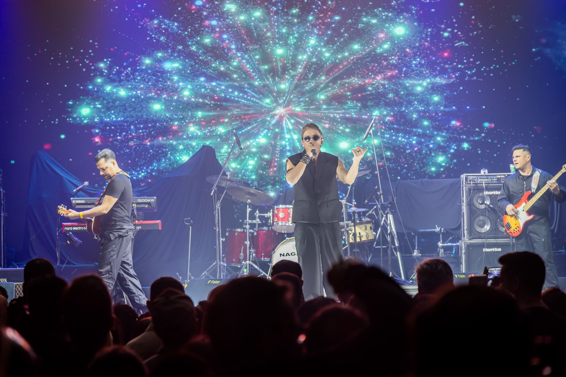 Uma banda se apresenta no palco diante de uma plateia, com uma exibição de luzes em formato de estrela projetada na tela.