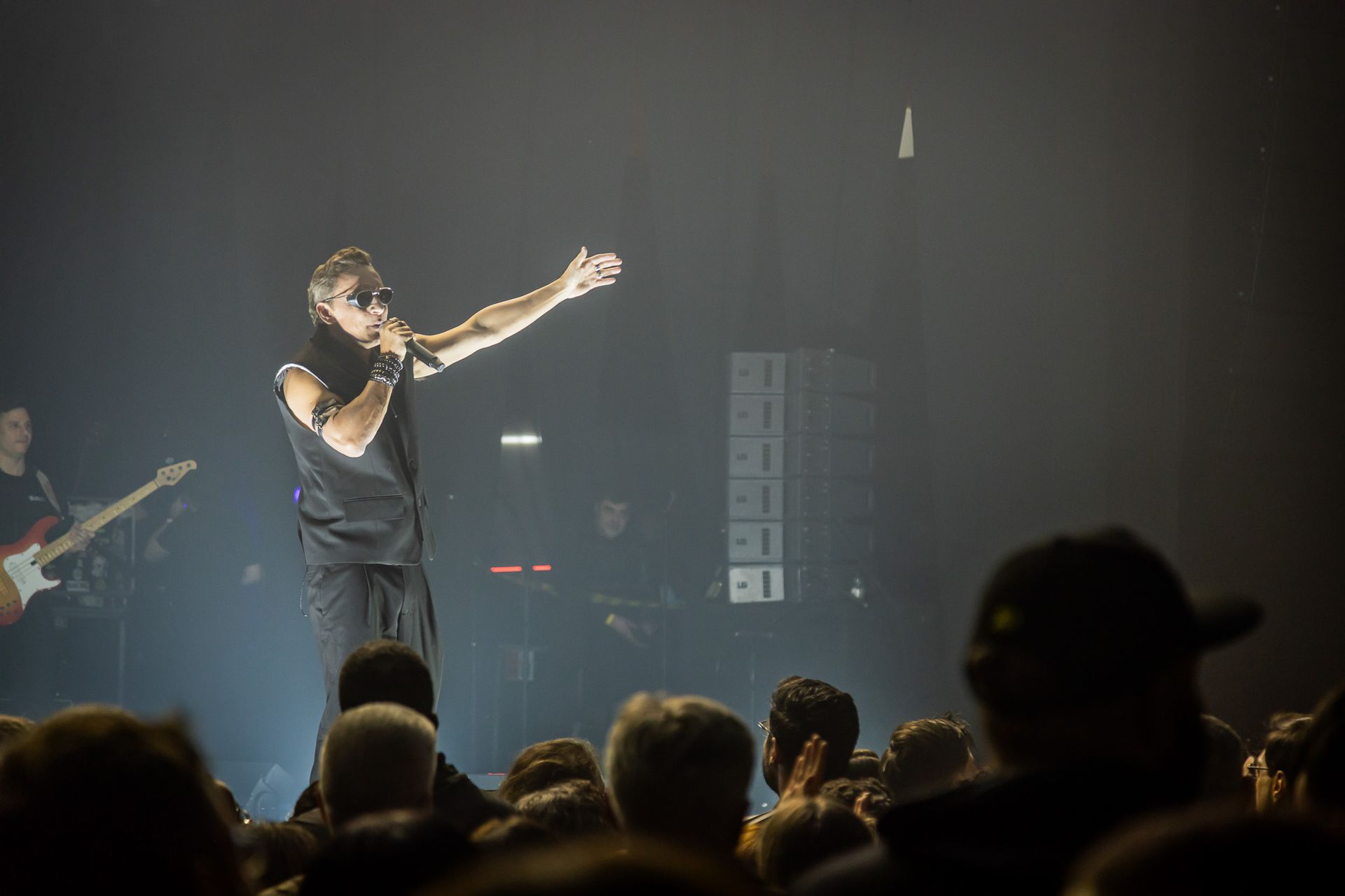 Um artista canta ao microfone, interagindo com o público em um palco de concertos com pouca luz.