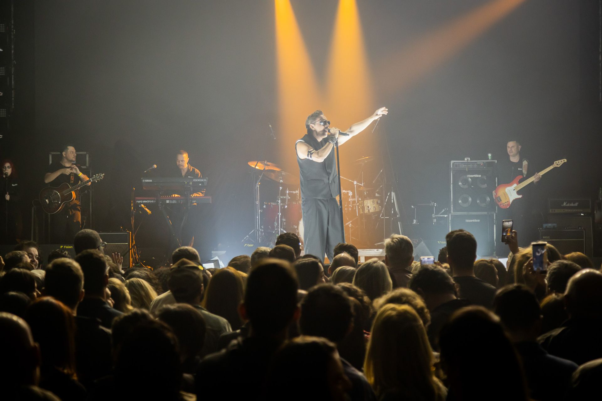Um artista cantando no palco, com o braço estendido, iluminado por holofotes diante de uma grande plateia.