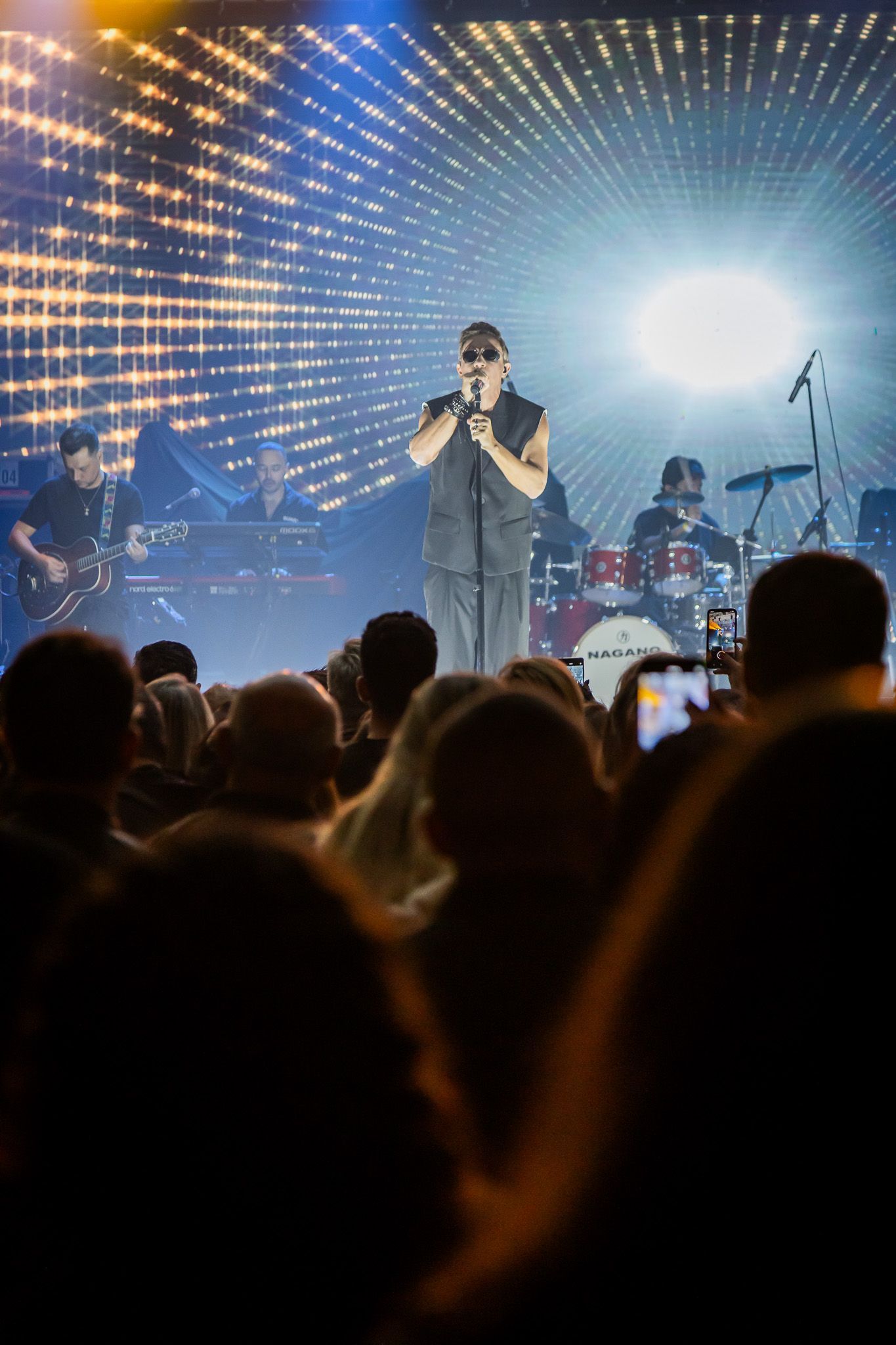 Um cantor se apresenta no palco com uma banda diante de uma plateia, emoldurada por um fundo de luzes brilhantes.