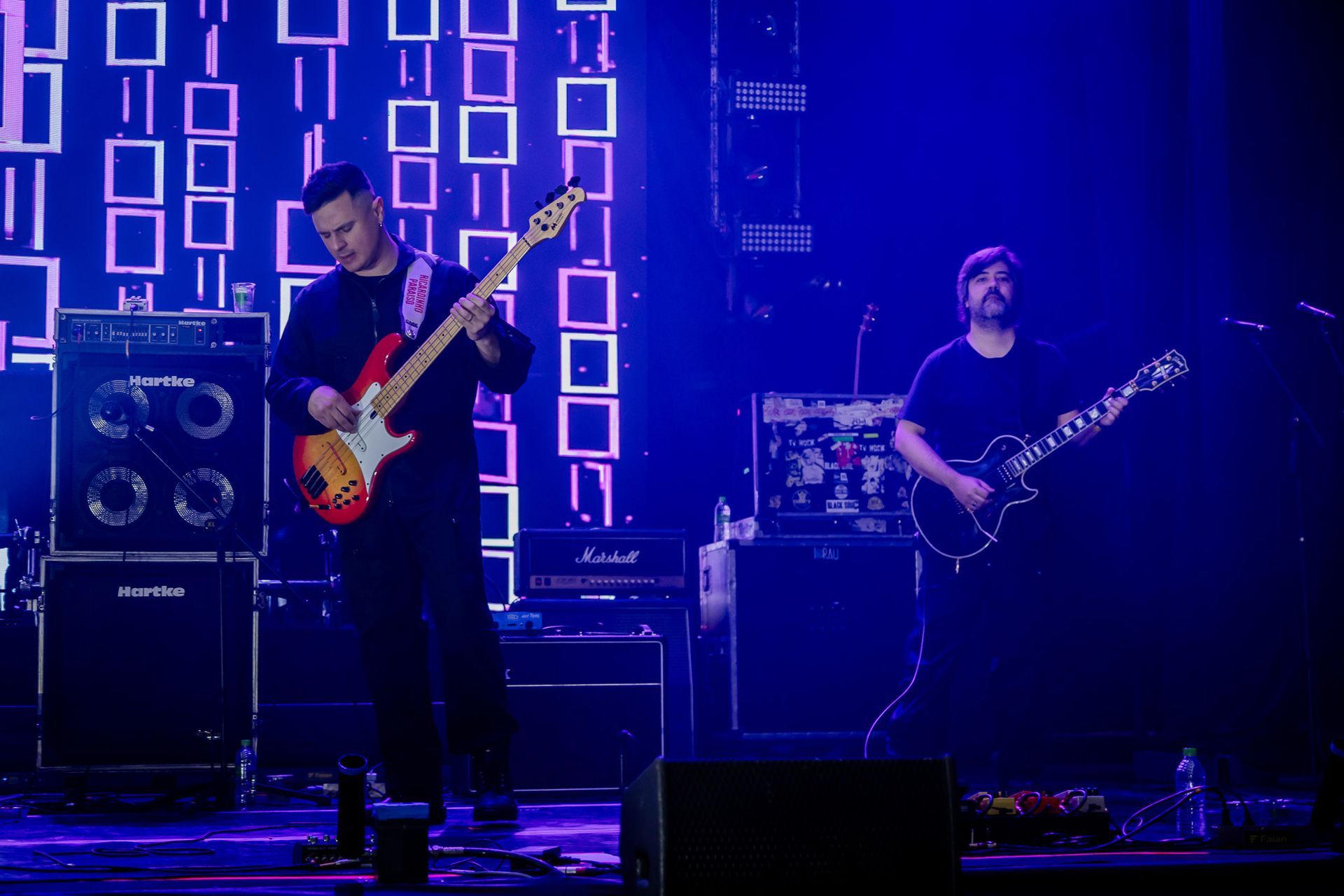 Dois músicos se apresentam no palco banhados por luz azul, um tocando um baixo vermelho e o outro uma guitarra elétrica.