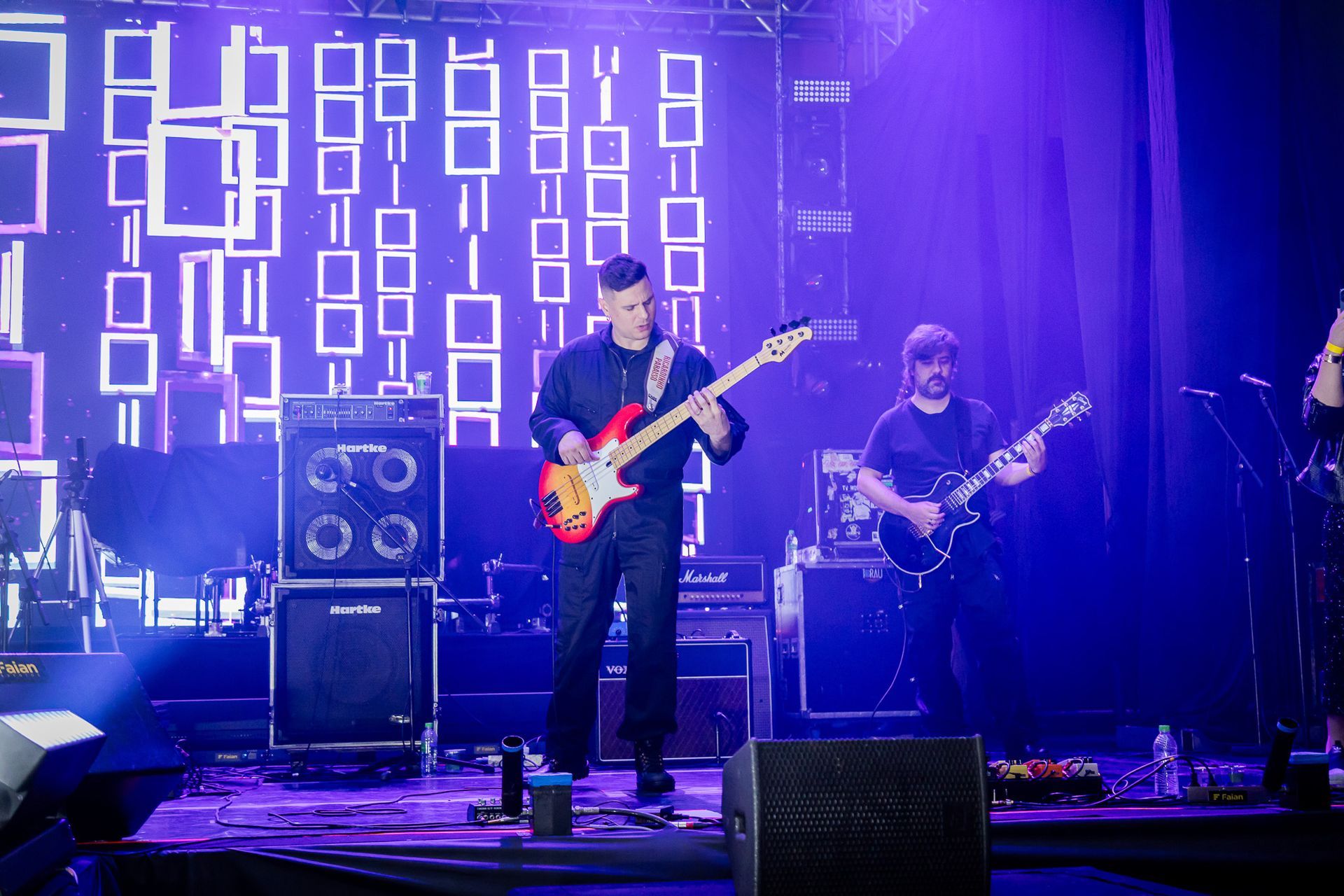 Uma banda se apresenta em um palco banhado por luz azul, tendo como pano de fundo padrões retangulares iluminados.
