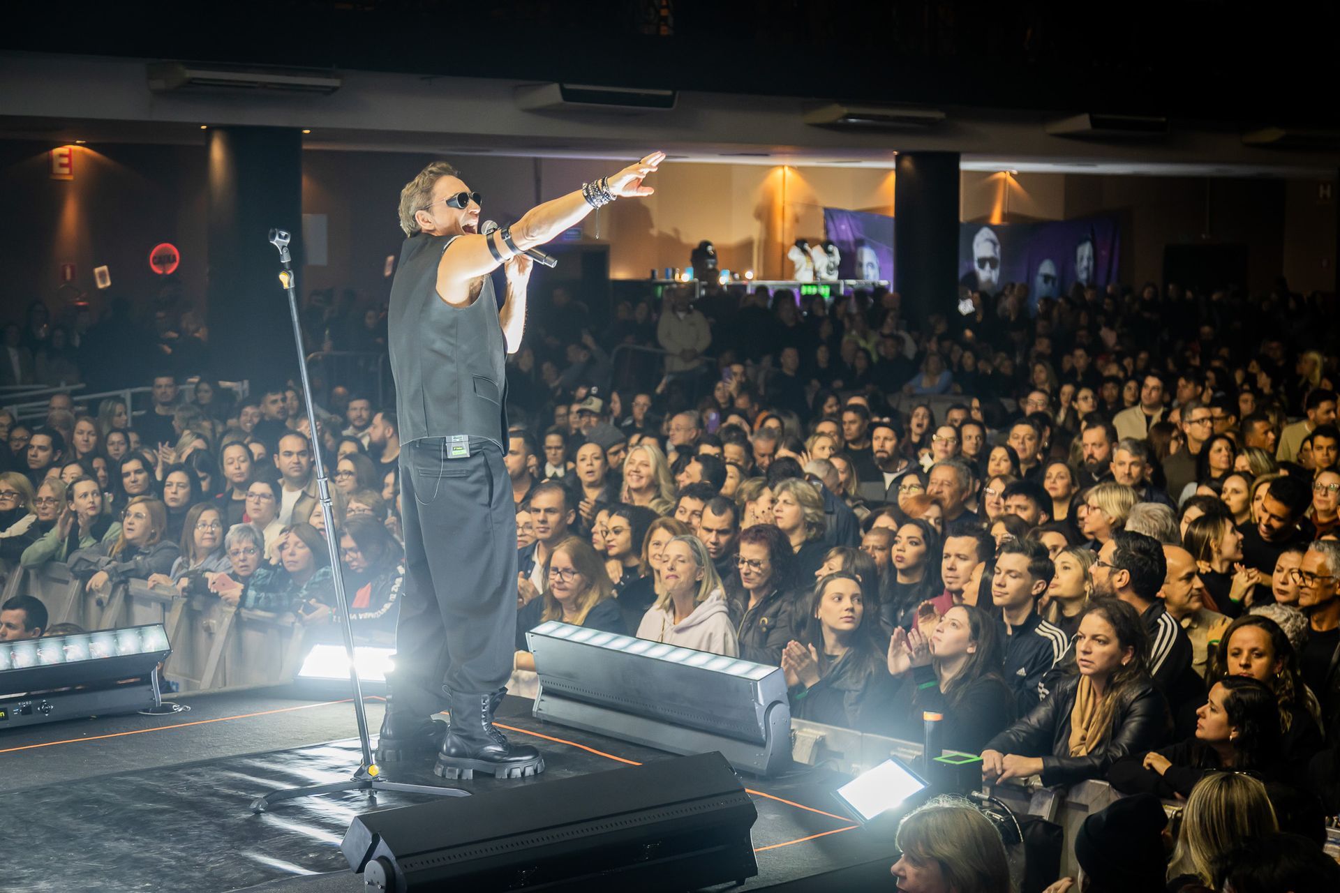 Um artista vestido de preto está no palco, gesticulando para uma grande plateia em um show com iluminação tênue.