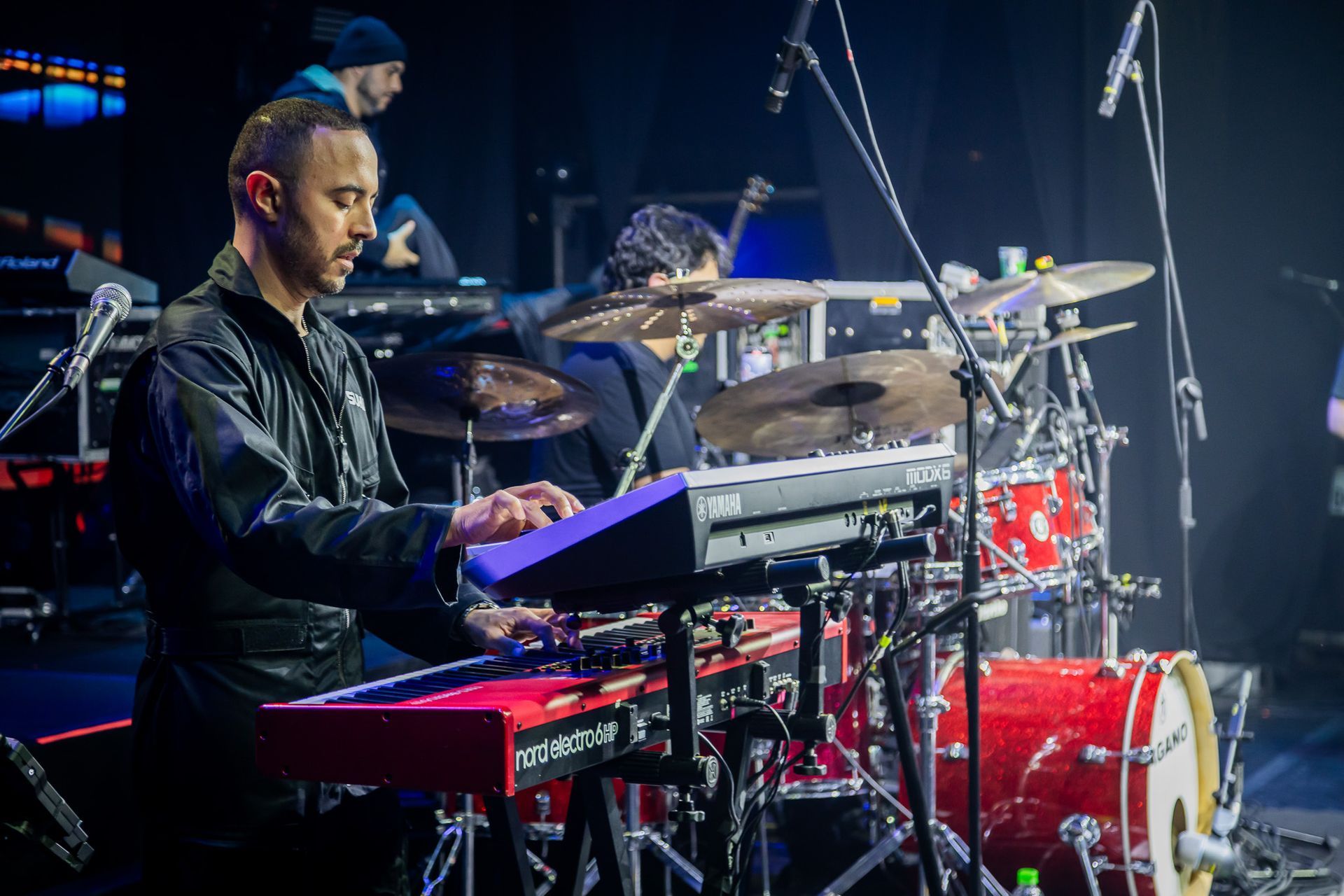 Um músico toca um teclado vermelho no palco, enquanto um baterista se apresenta ao fundo, em meio à iluminação