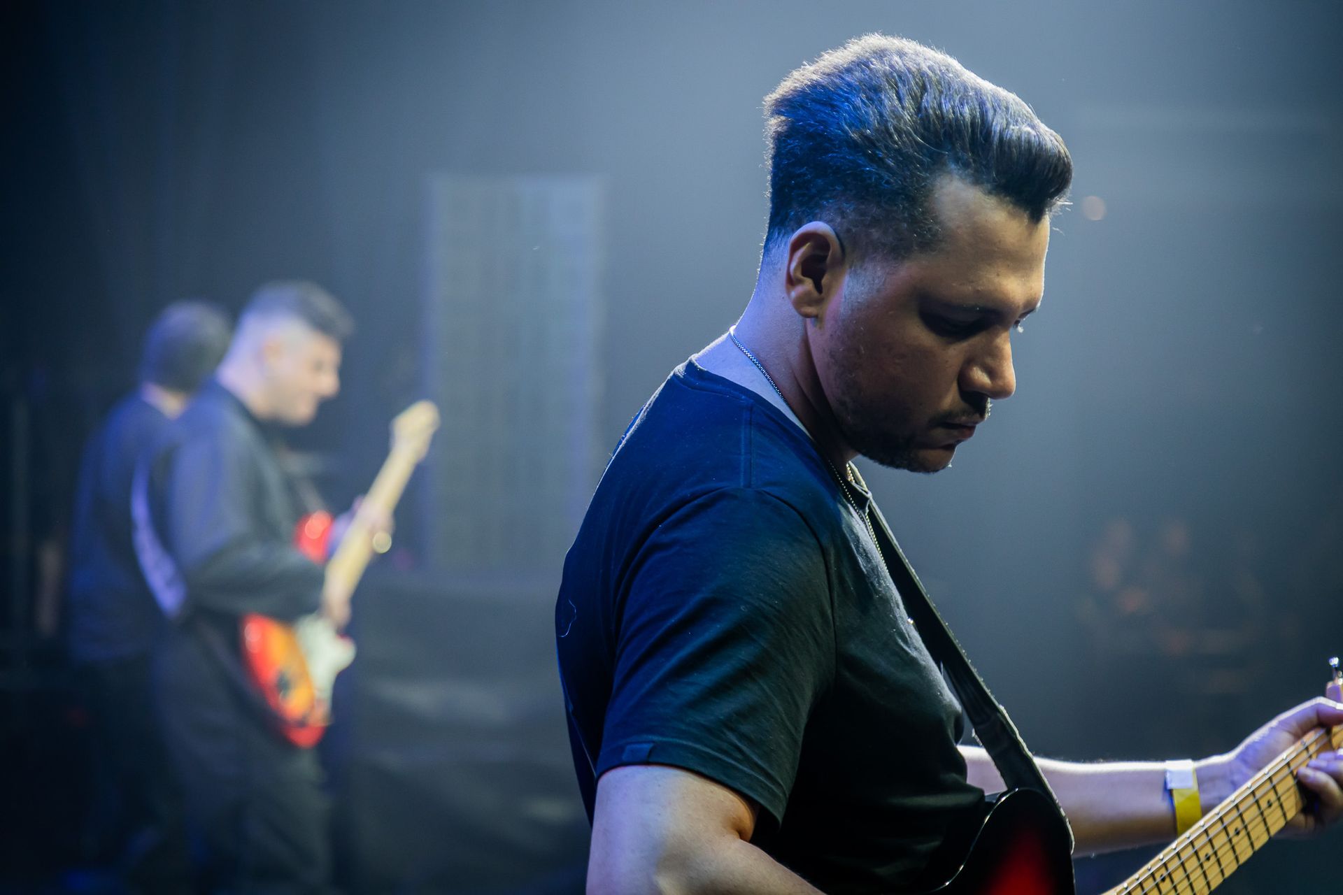 Dois músicos de camisa escura tocam guitarras elétricas em um palco com iluminação tênue e atmosférica.