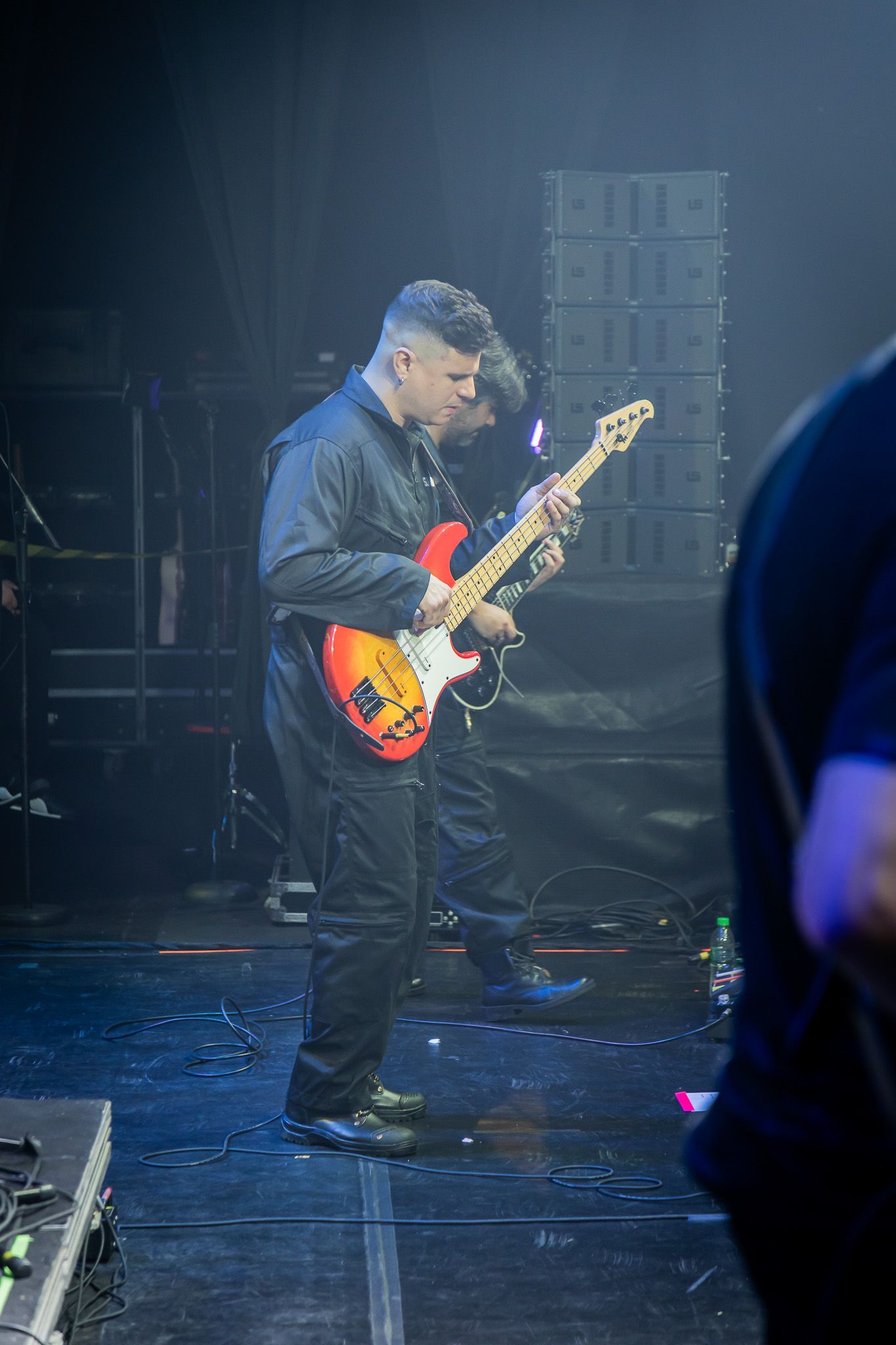 Um músico toca um baixo elétrico sunburst em um palco de concerto com pouca luz.