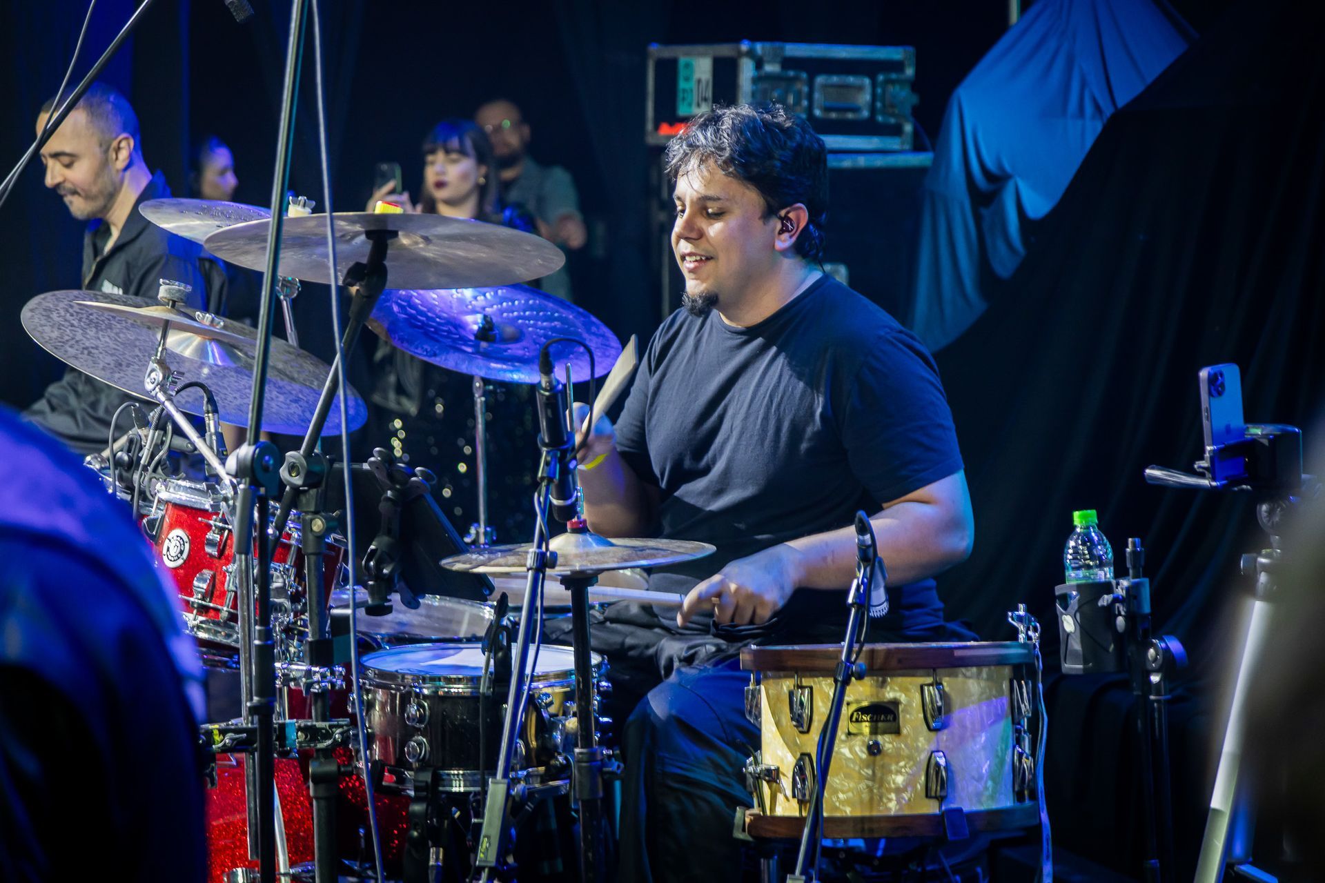 Um baterista sorridente, vestindo uma camiseta preta, toca bateria em um palco com pouca luz.
