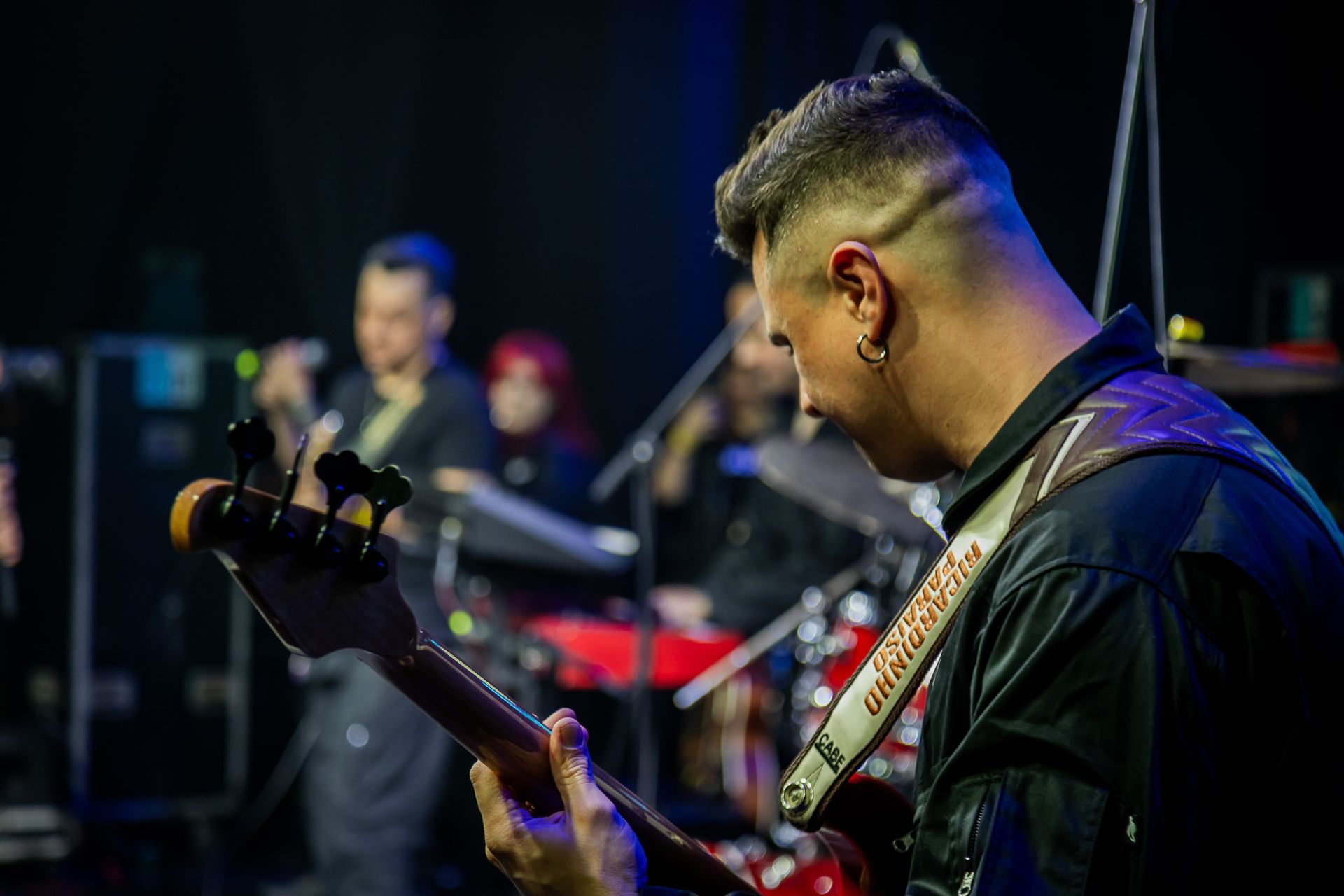 Um músico toca baixo em um palco escuro e pouco iluminado, com os membros da banda desfocados ao fundo.