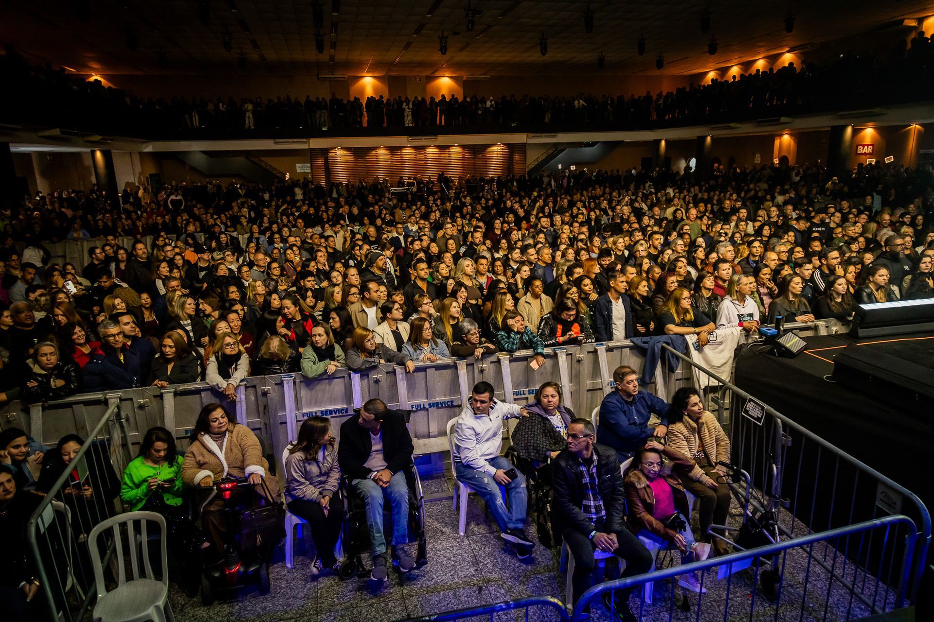 Um grande espaço fechado para concertos, lotado de público, com pessoas sentadas em frente a um palco atrás de uma barreira.
