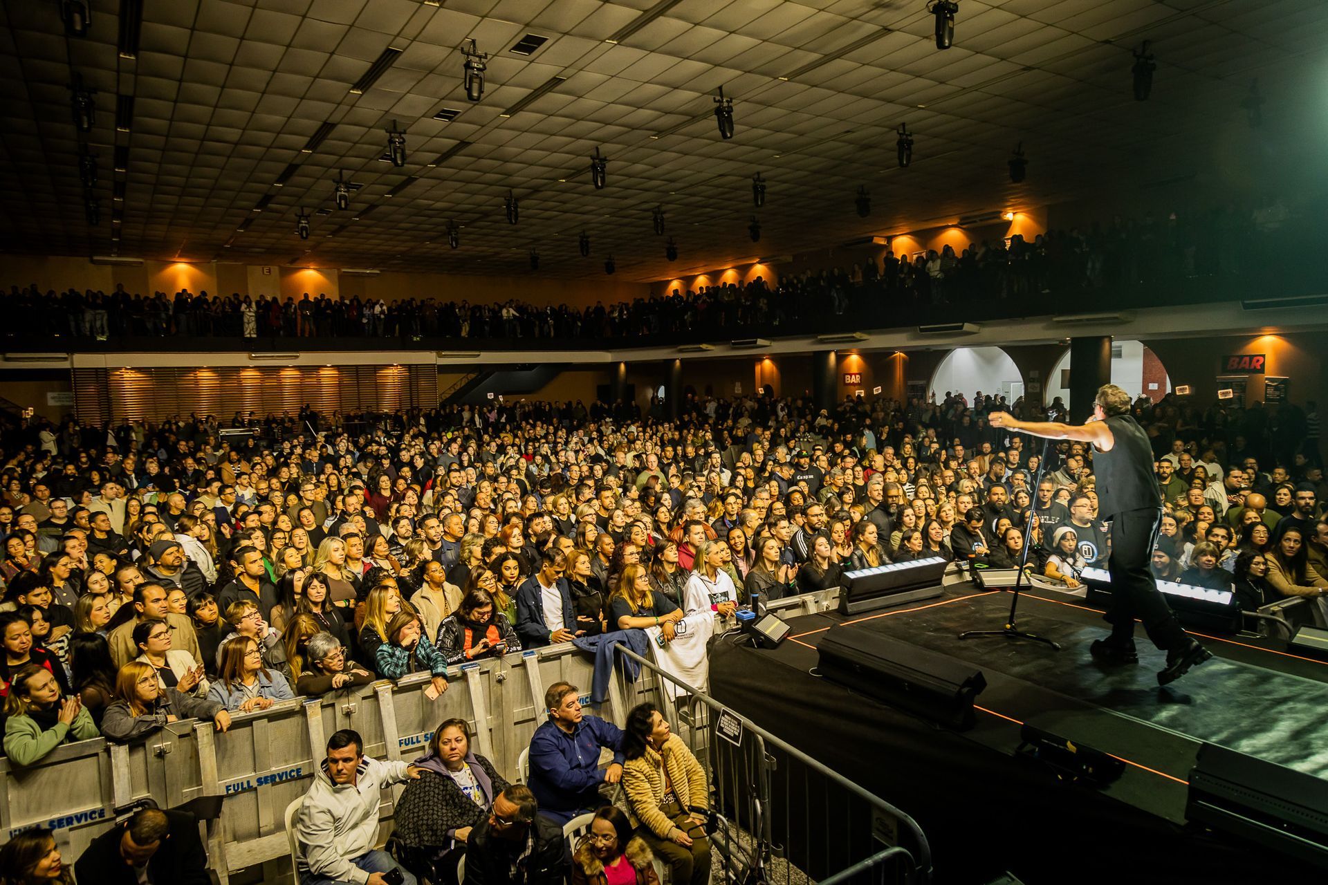 Um artista no palco gesticula em direção a uma grande plateia sentada em uma sala de concertos com pouca luz.