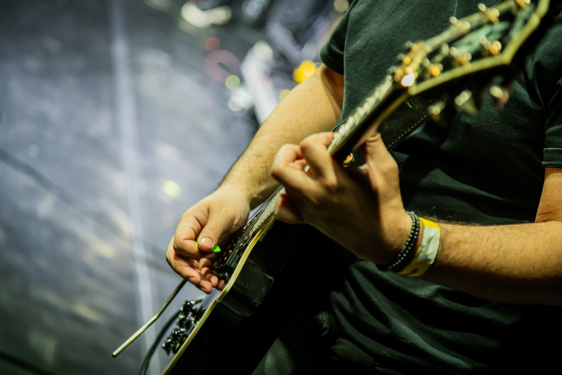 Um músico de camisa preta toca uma guitarra elétrica com alavanca de vibrato, segurando uma palheta verde sobre as cordas.