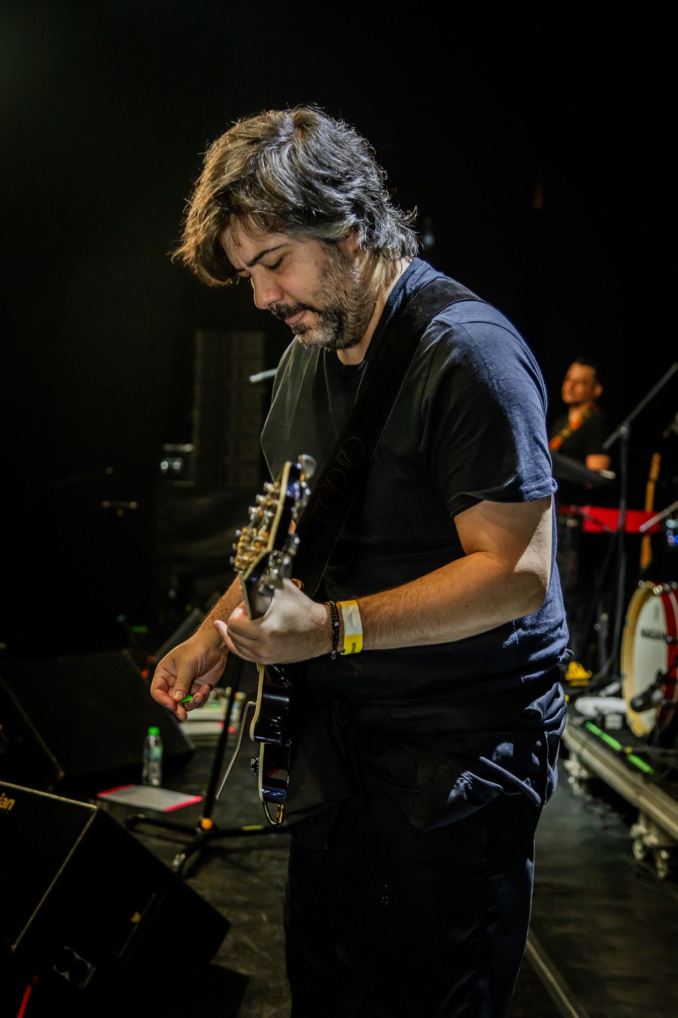Um músico de cabelos longos e escuros toca guitarra elétrica em um palco escuro, concentrado em suas mãos.