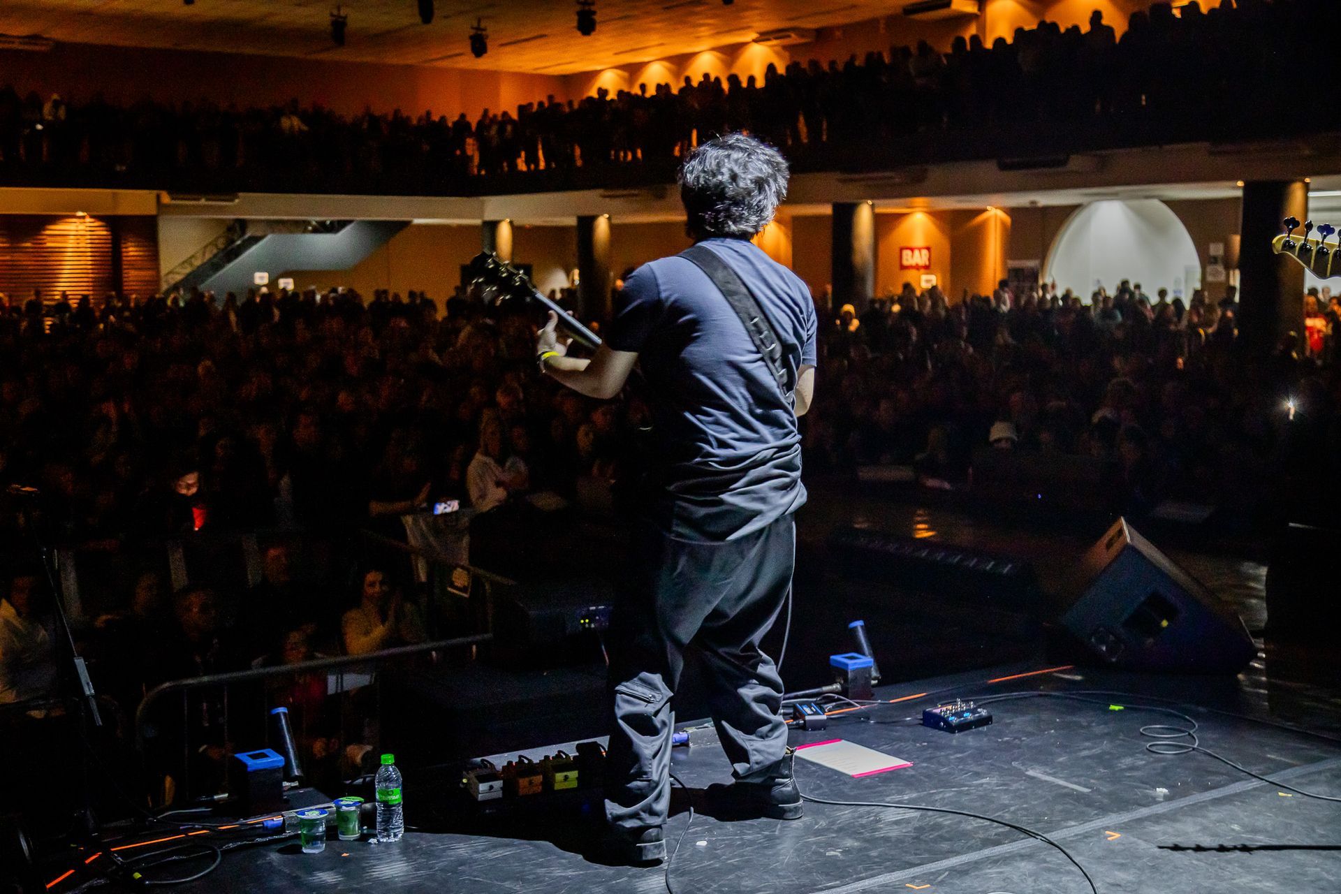 Um músico toca guitarra em um palco de frente para uma grande plateia, em uma casa de shows escura.