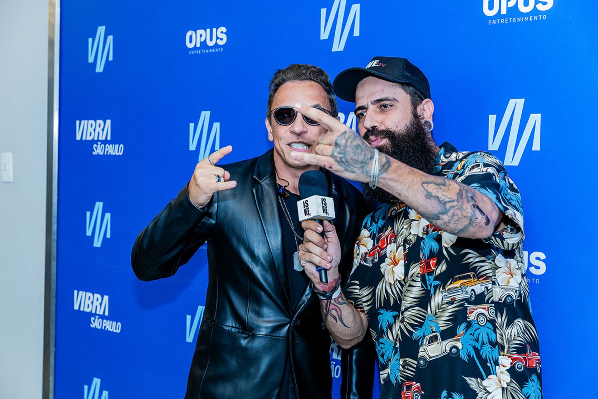 Dois homens posam em frente a um fundo azul com a marca da empresa, um deles segurando um microfone.