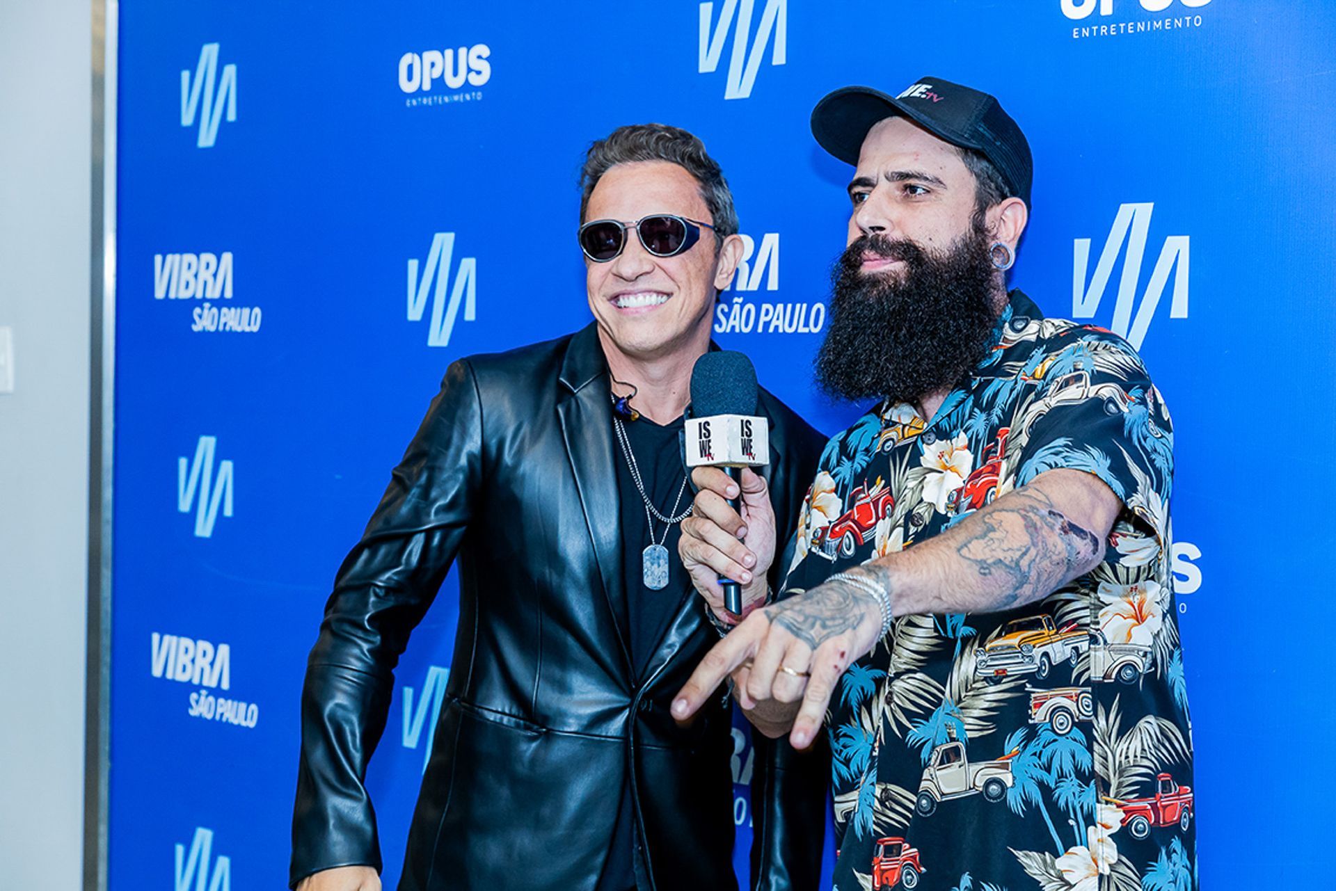 Um homem de jaqueta de couro e óculos escuros segura um microfone em direção a um homem barbudo com camisa tropical.