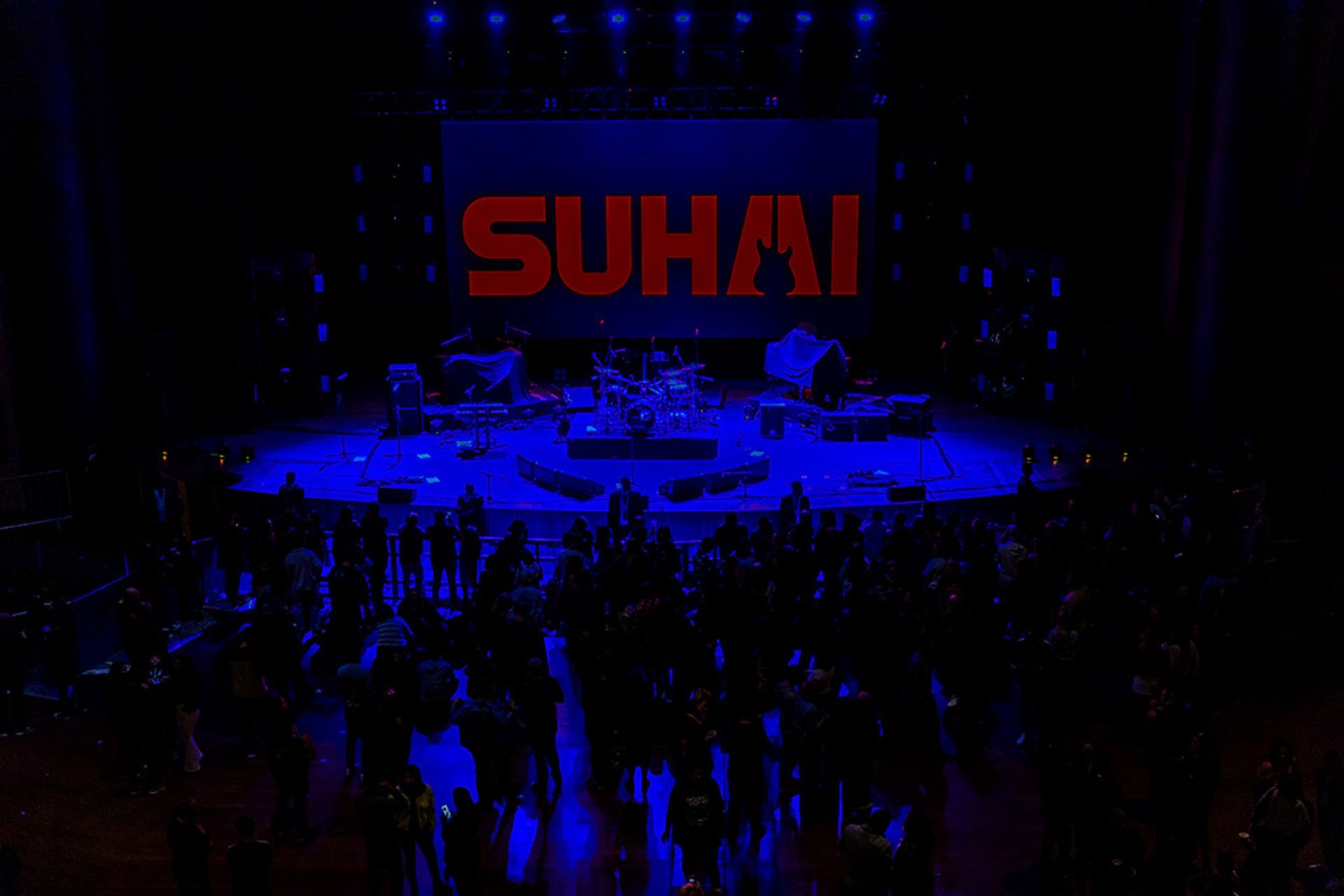Um palco de concertos banhado em luz azul, com um grande telão exibindo o texto vermelho 