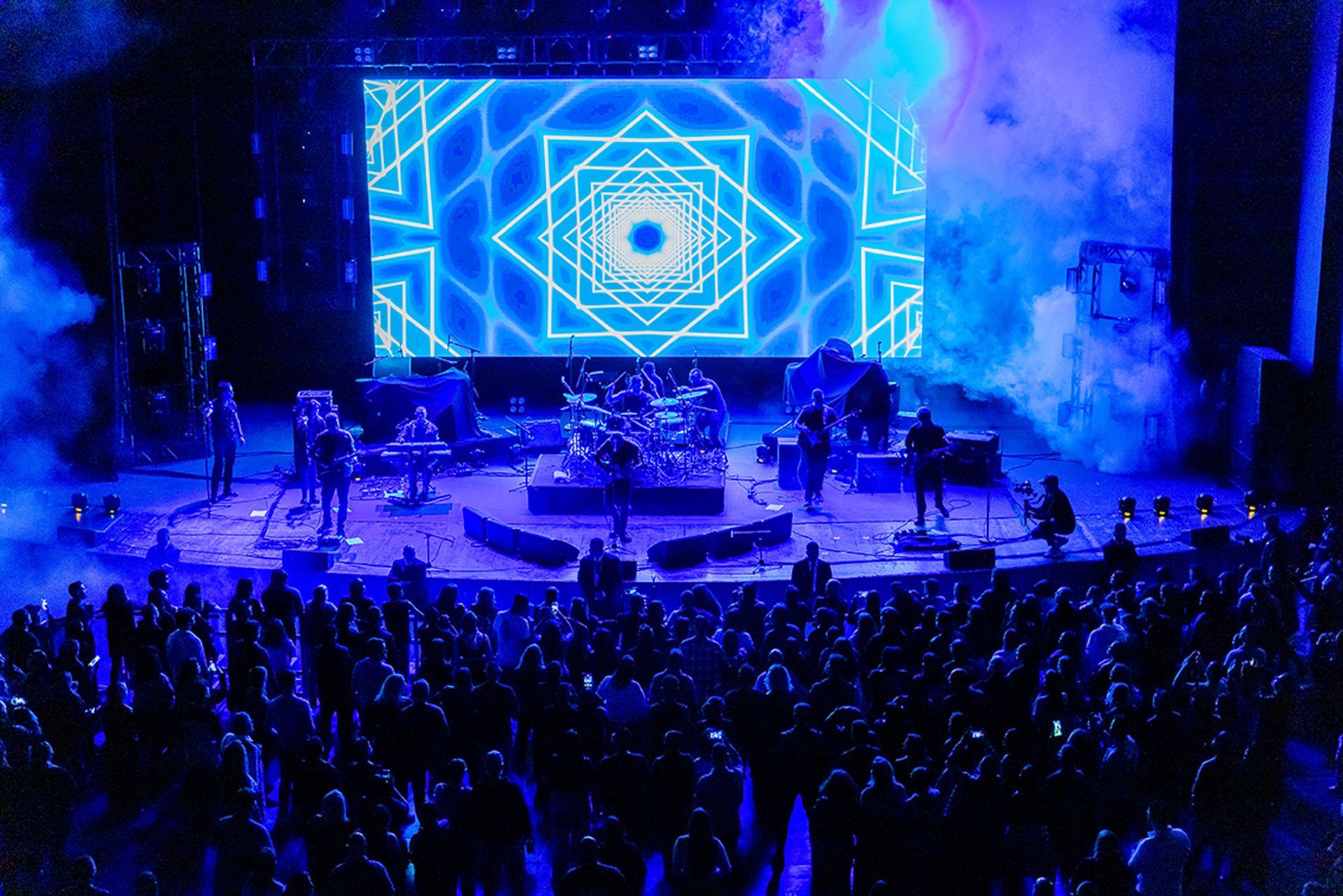 Uma banda se apresenta em um palco iluminado por luzes azuis e fumaça, com um padrão geométrico em uma tela e uma plateia.