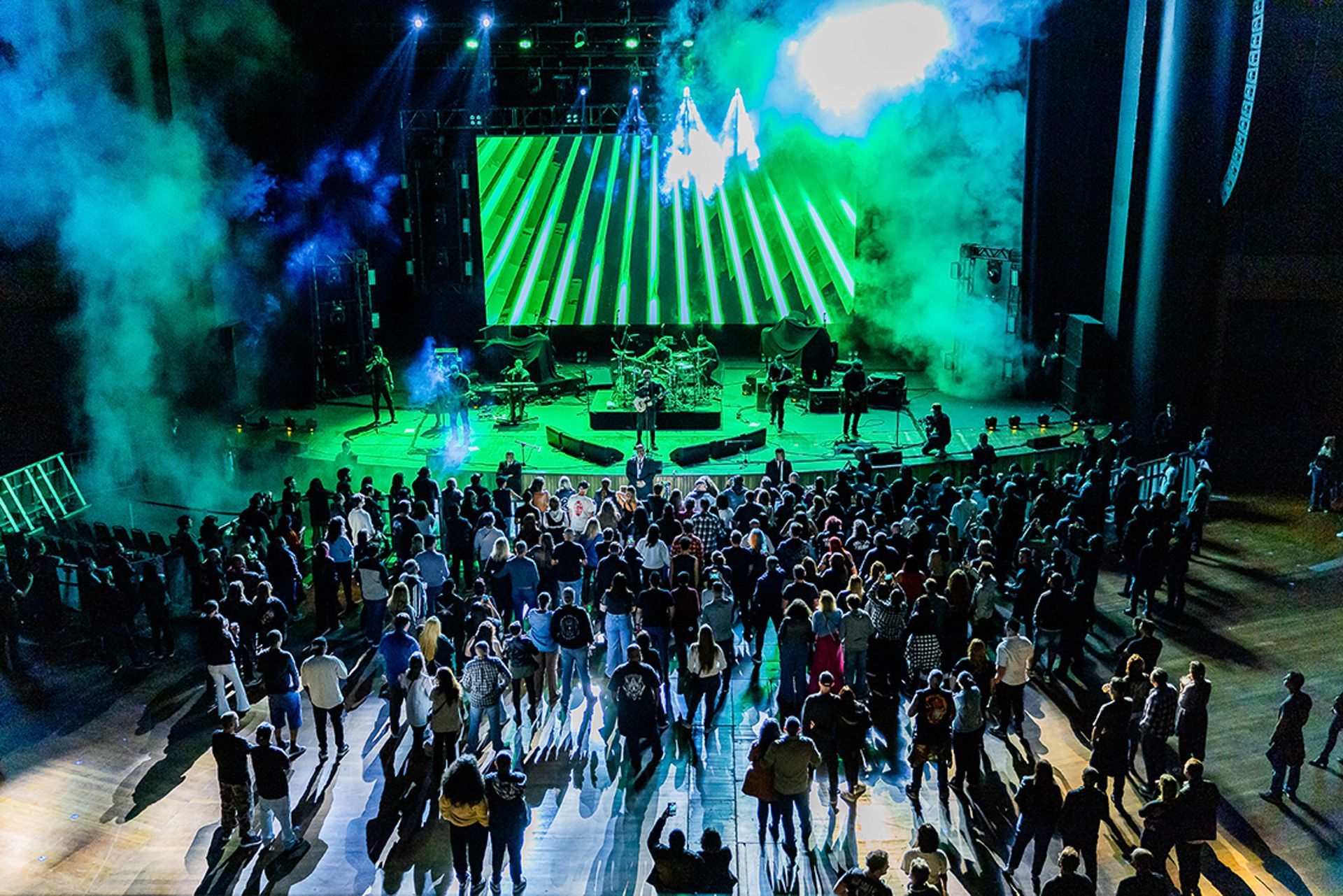 Vista de cima de um concerto com um palco verde brilhante, membros da banda e uma multidão em silhueta no auditório escuro.
