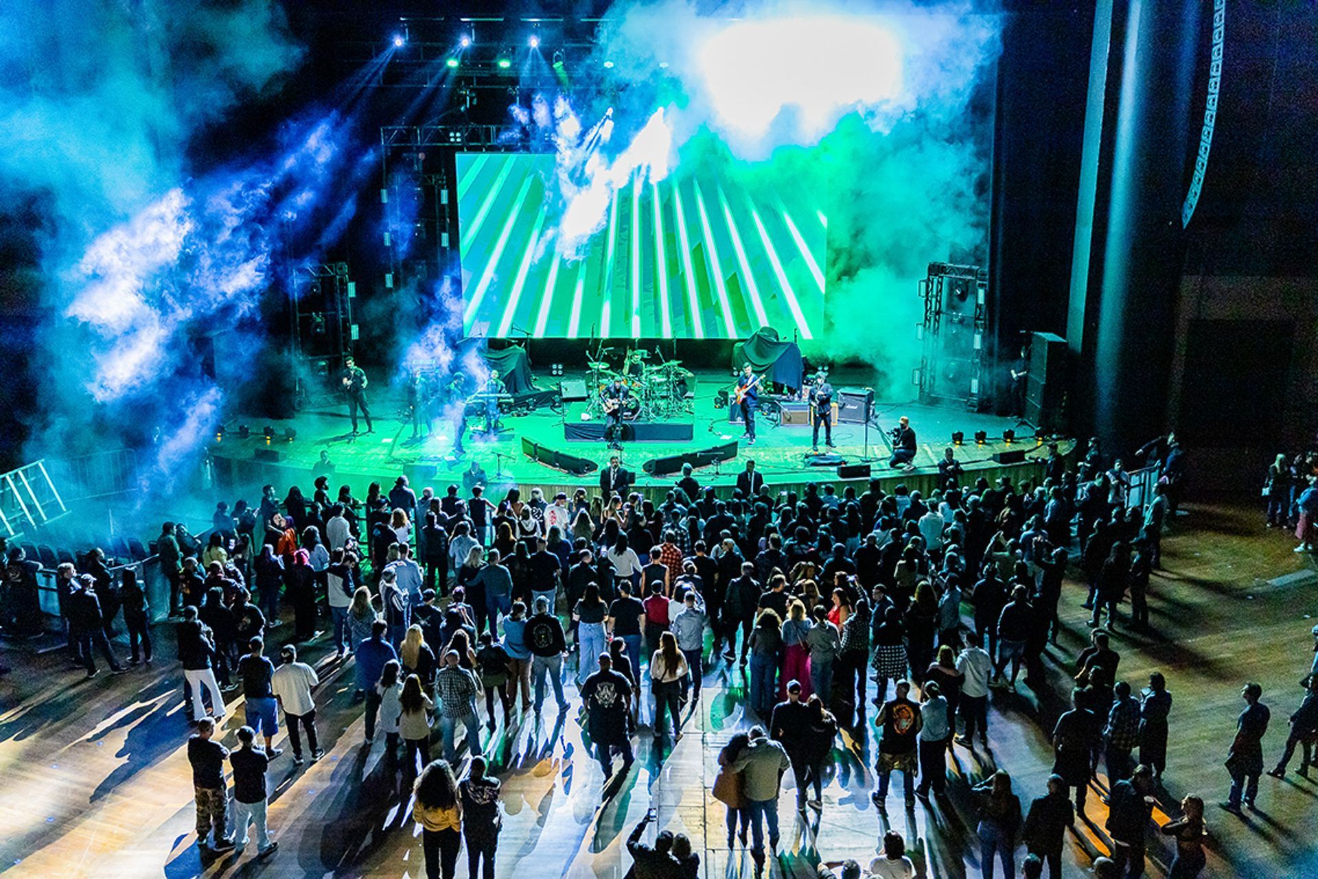 Uma visão panorâmica de uma multidão em um show ao ar livre, com um palco bem iluminado, tela verde e fumaça azul acima.