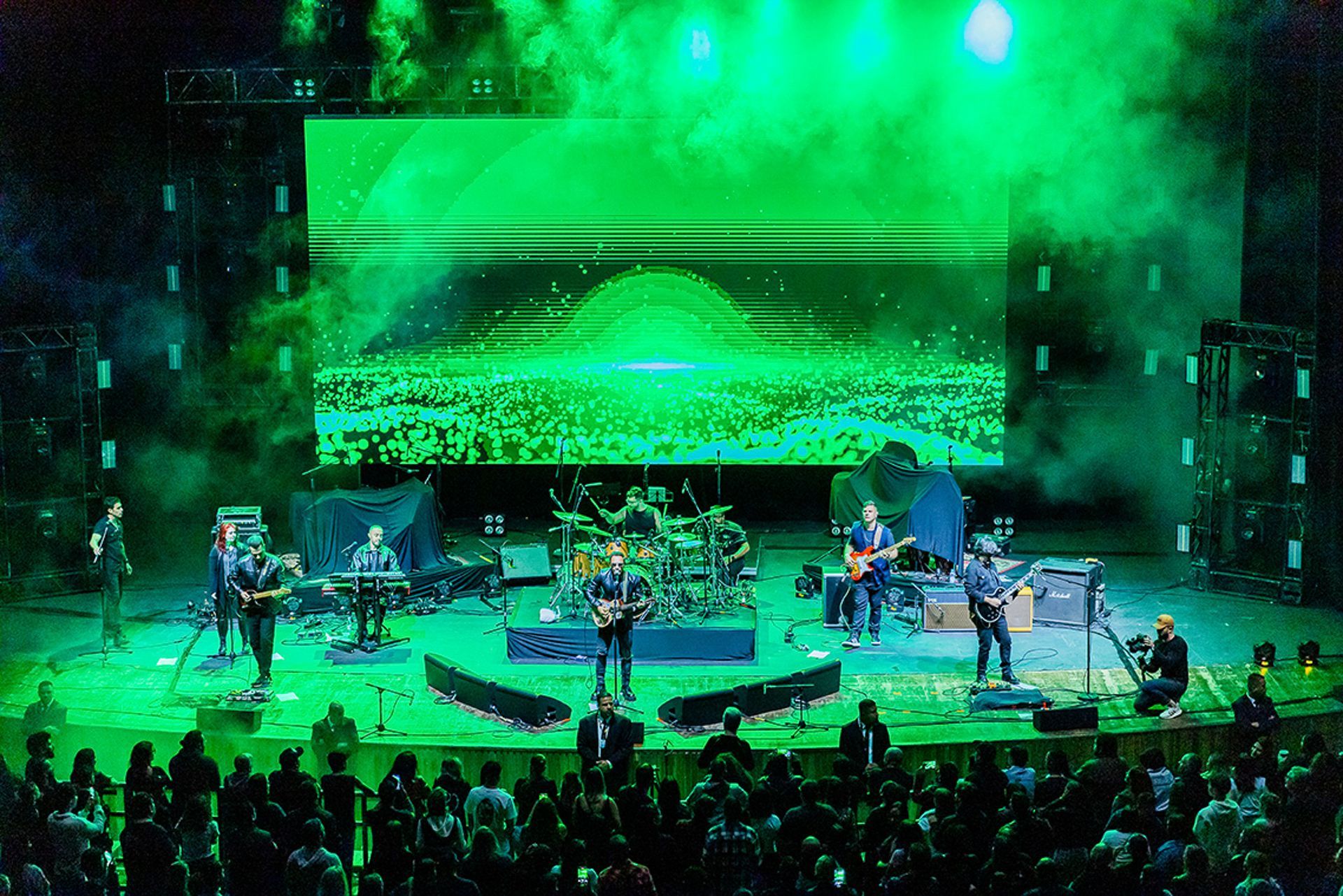 Uma banda se apresenta em um palco verde bem iluminado, em frente a um telão e uma plateia, em uma casa de shows fechada.