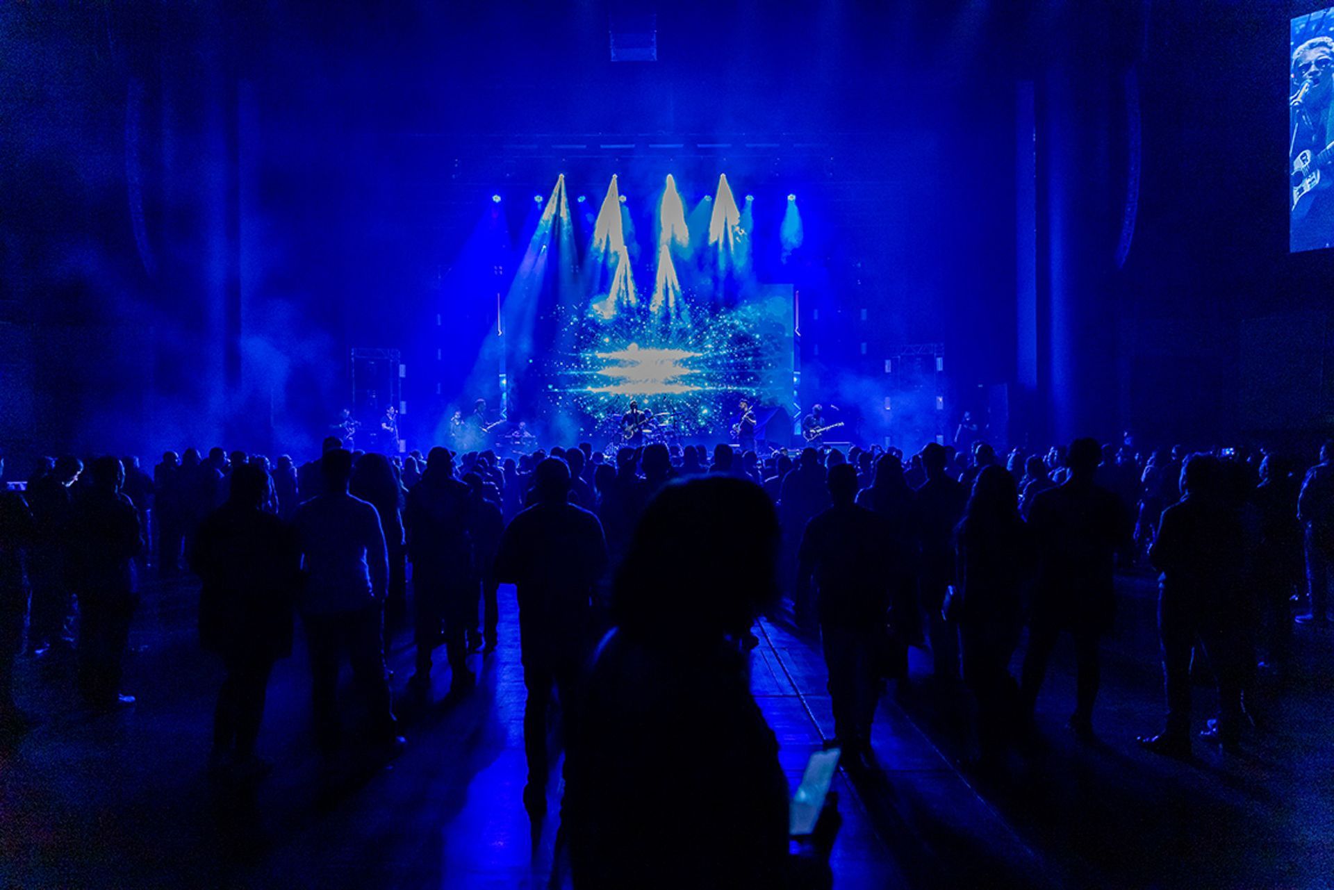 Uma multidão se destaca contra um palco azul brilhante durante um concerto, com feixes de luz incidindo sobre os artistas.