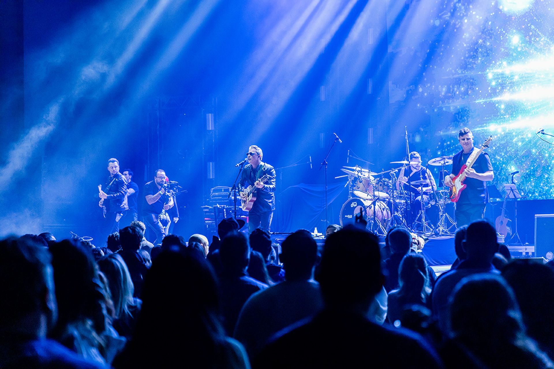 Uma banda se apresenta no palco sob fortes holofotes azuis diante de uma plateia em uma sala de concertos.