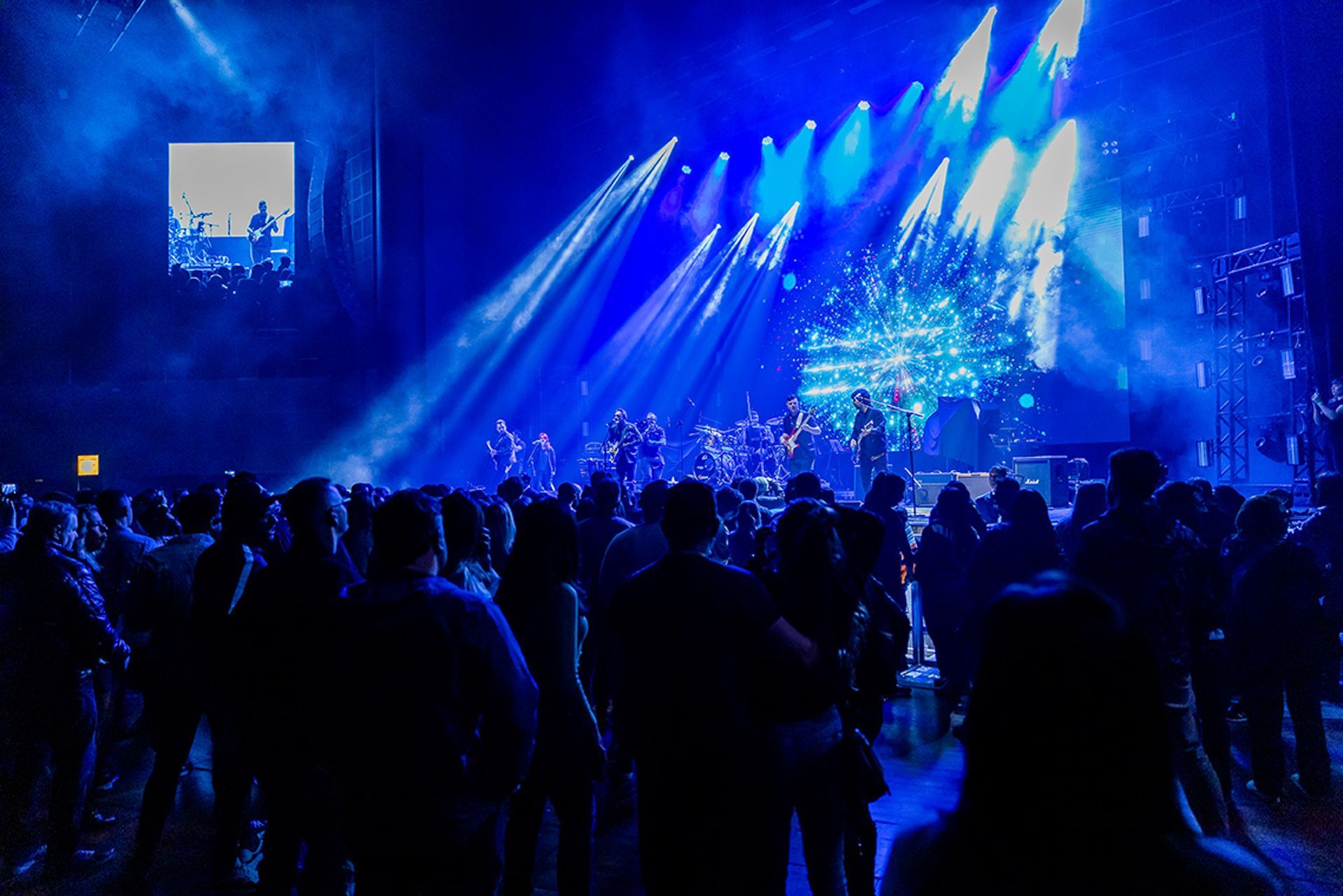 Uma multidão assiste a um concerto com iluminação de palco azul, feixes de luz dinâmicos e um telão exibindo silhuetas.