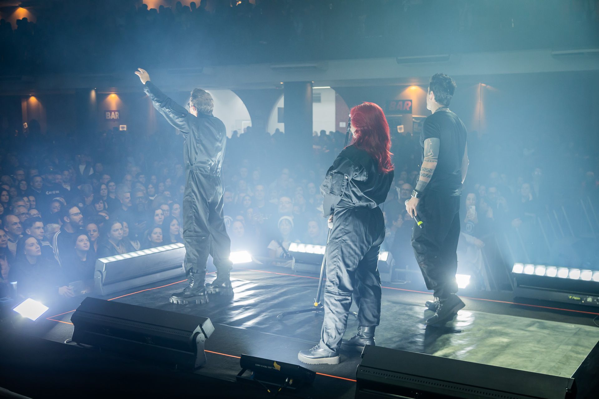 Três artistas se apresentam em um palco pouco iluminado diante de uma grande plateia, banhada por holofotes e uma névoa.