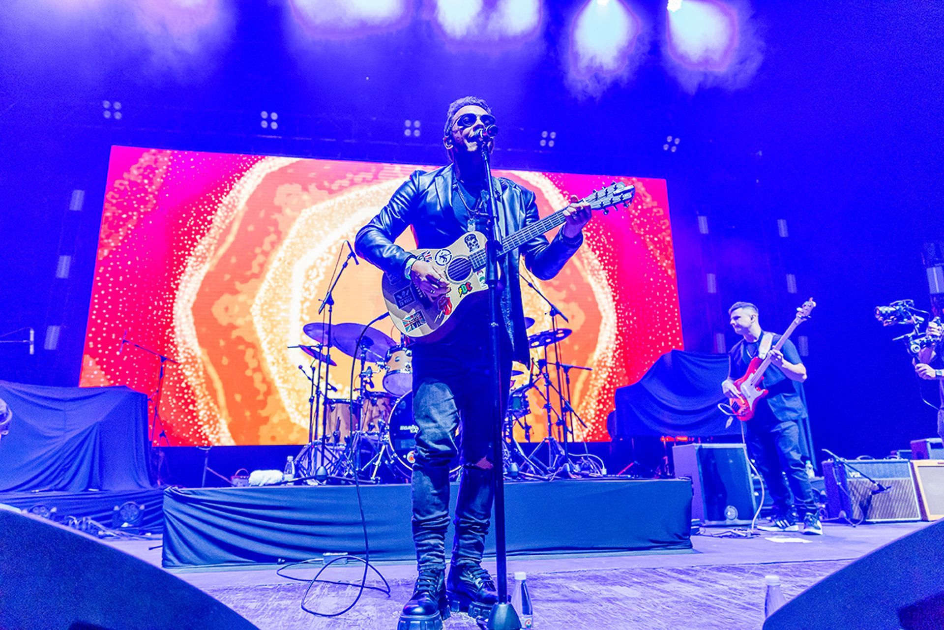 Um artista com um violão está no centro do palco sob luzes azuis, diante de uma tela com um padrão laranja vibrante.